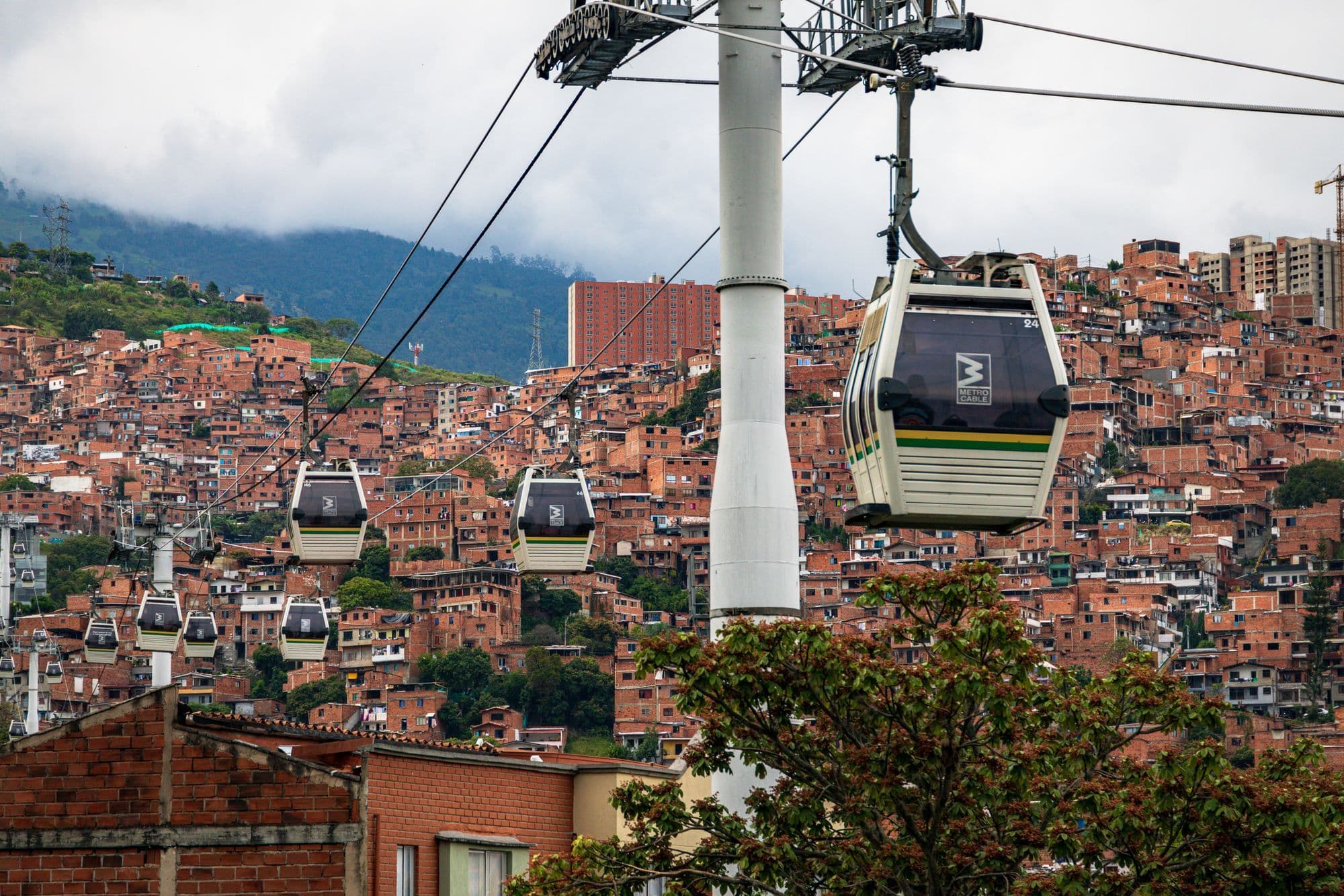 Metrocable gondolas hanging from cables above a hillside of red-brick houses in Medellín, with green hills behind.