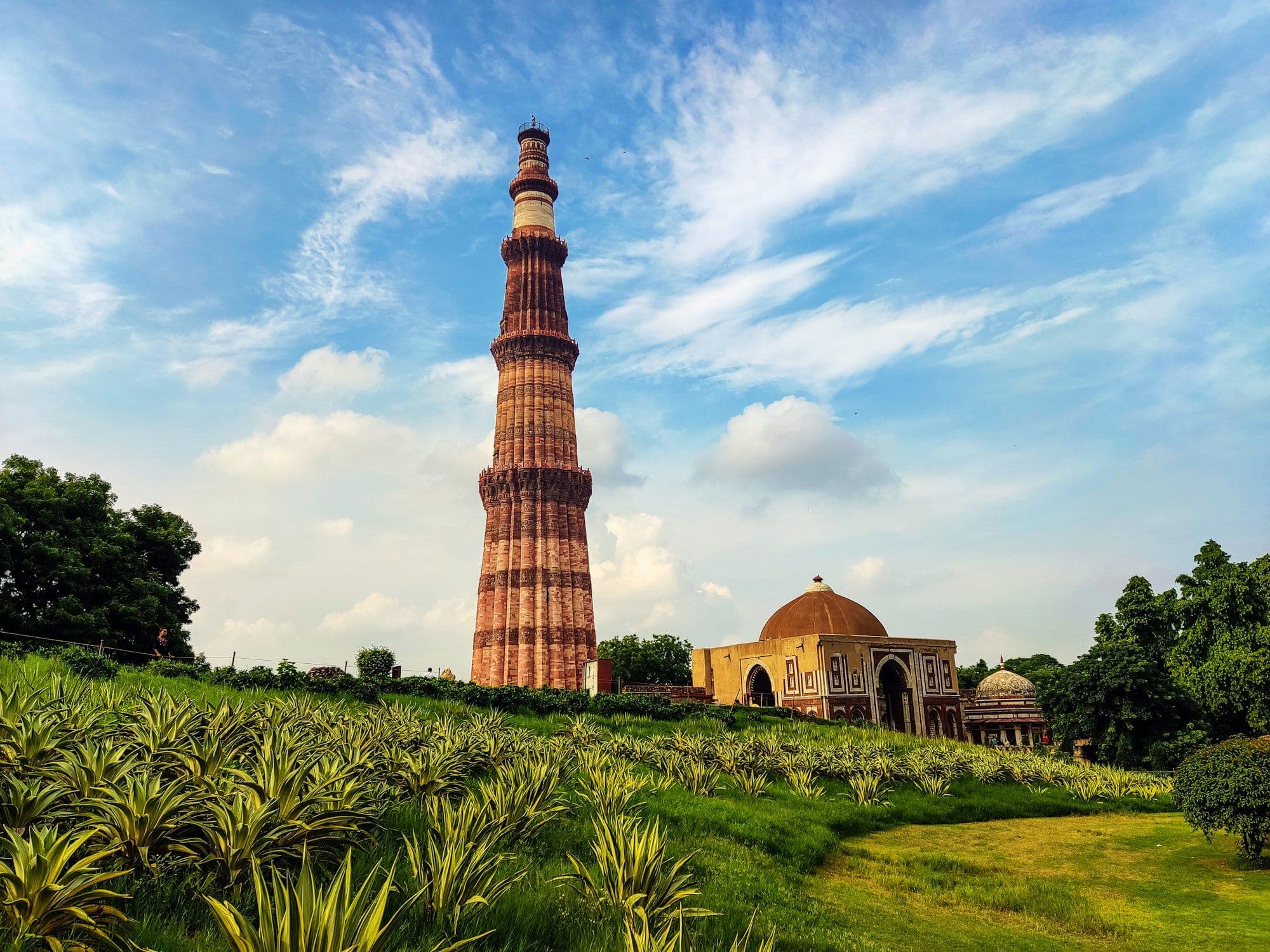 The fluted red sandstone tower of the Qutub Minar rising above lawns and a domed gateway under a blue sky.