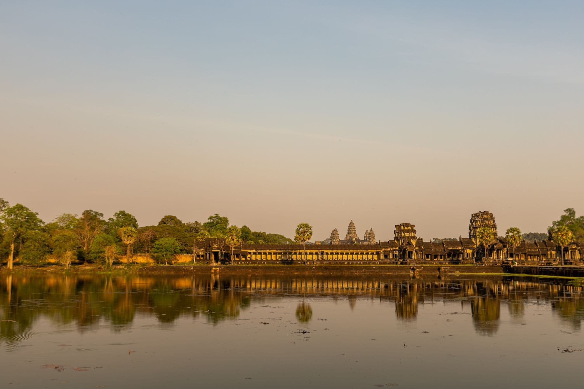 The five towers of Angkor Wat reflected in a still pond at dawn, under a pale orange sky.
