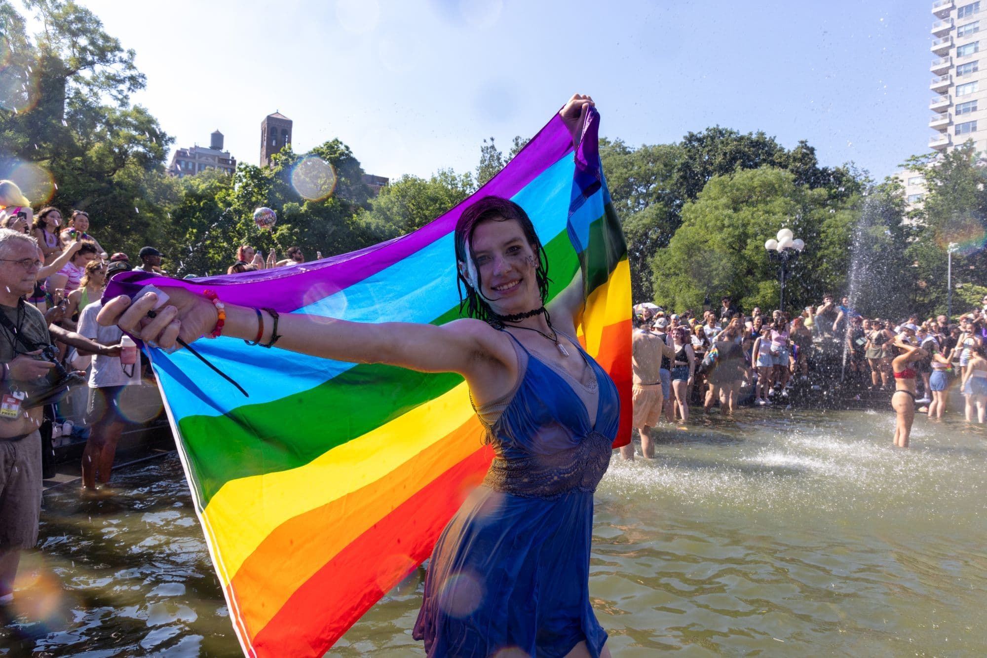 A soaked marcher in a blue dress holding a rainbow flag open while standing in a park fountain during New York City Pride, crowds cooling off behind.