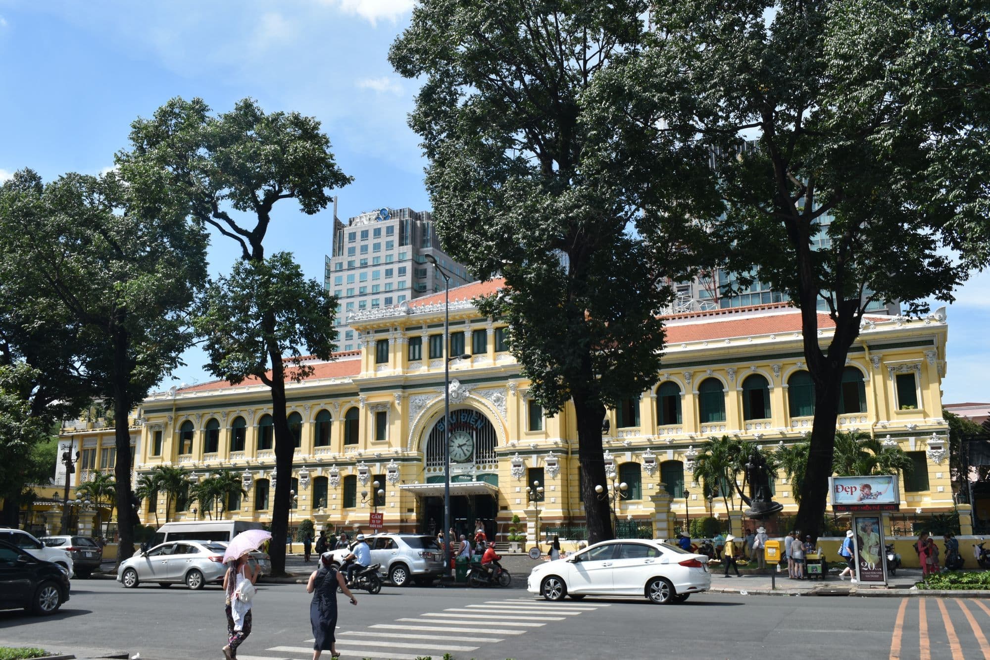 The yellow colonial facade of Saigon Central Post Office behind tall trees, with cars and pedestrians on the square in front.