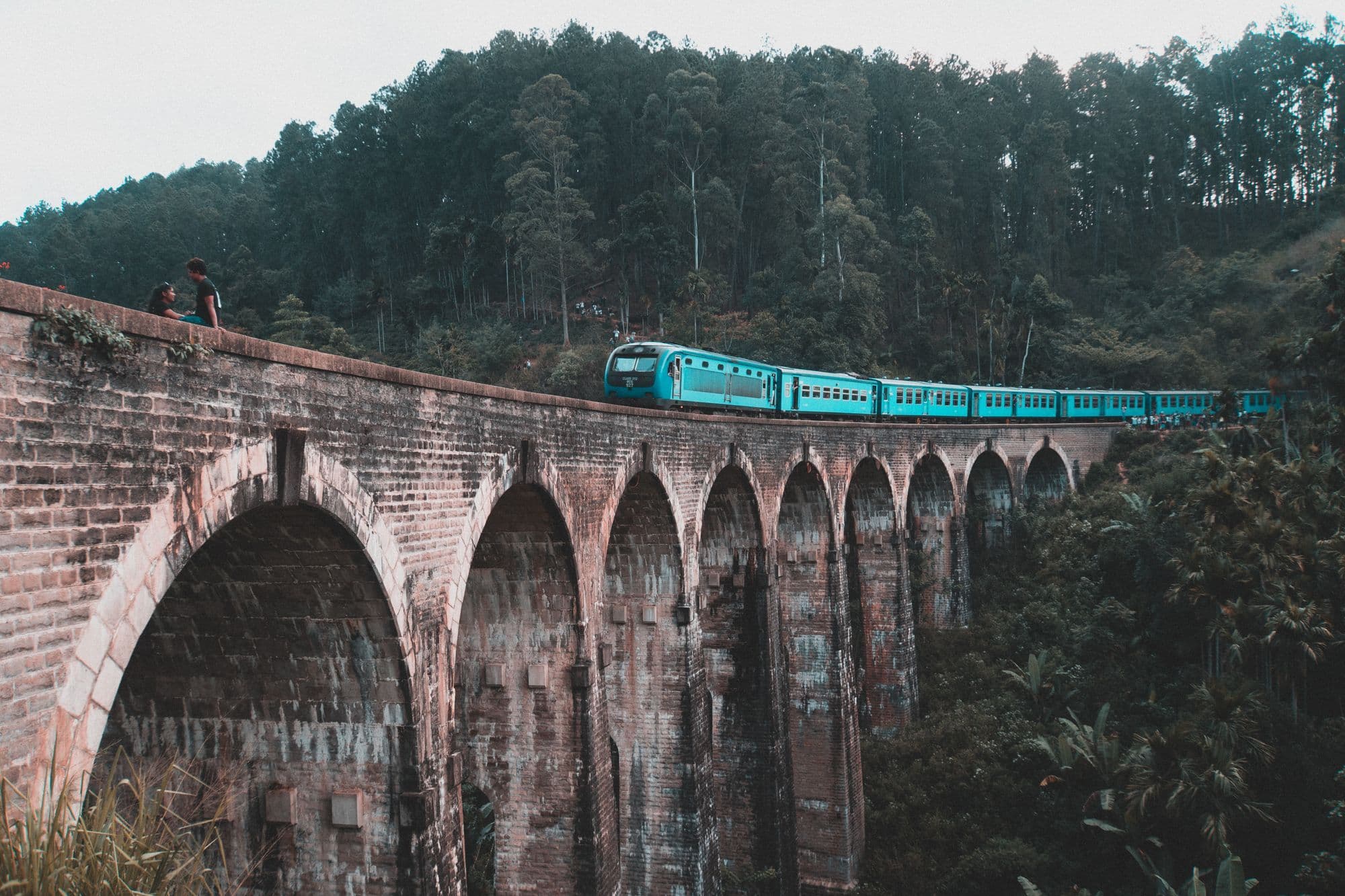 A blue train crossing the curved brick Nine Arch Bridge high above a forested valley near Ella.