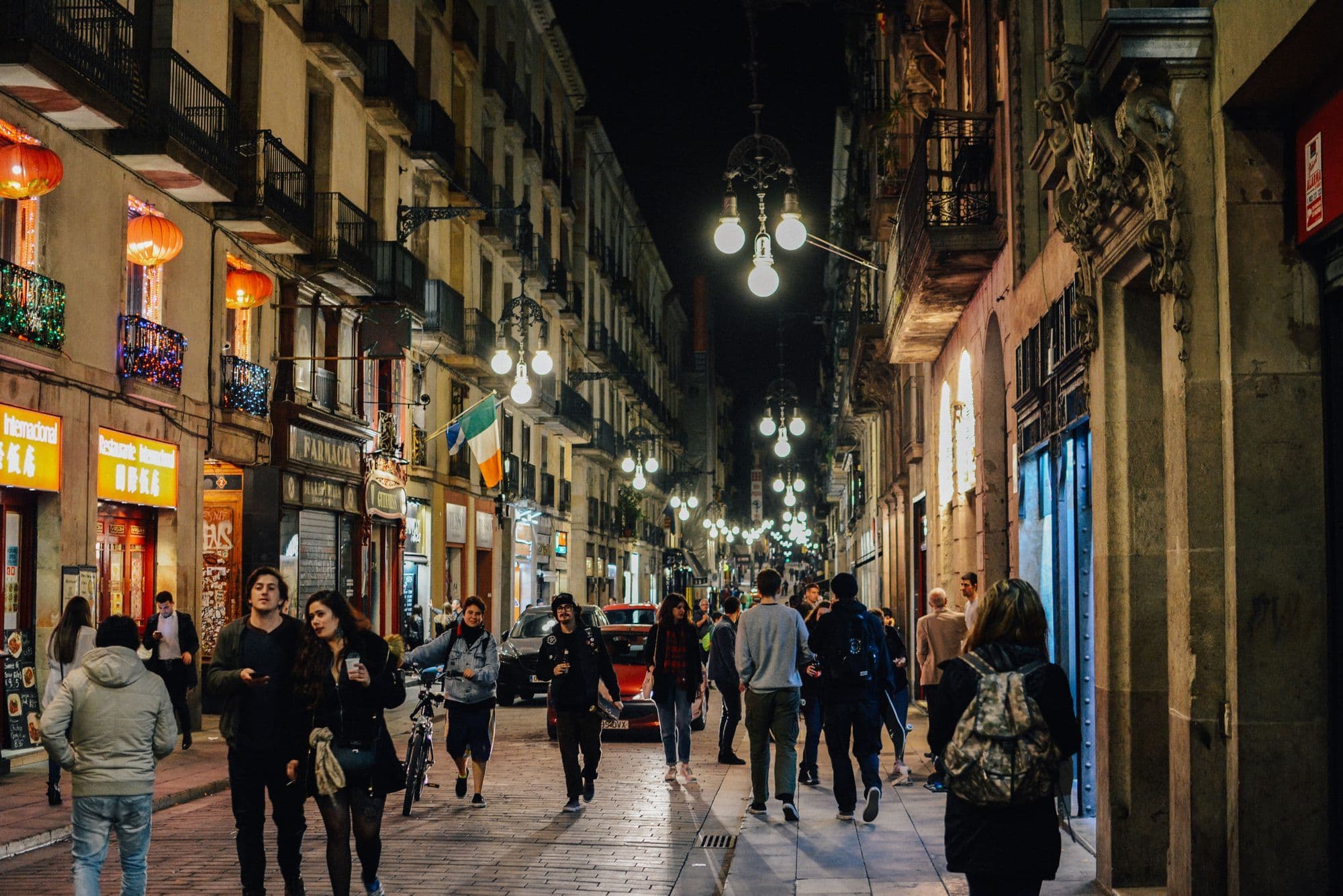 A narrow old-town street in Barcelona at night, lit by hanging lamps, with people walking between shopfronts.