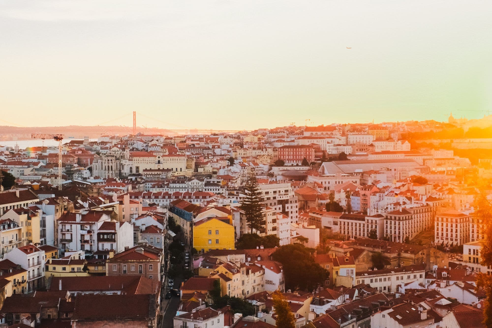 Lisbon's terracotta rooftops and pale facades glowing at sunset, with a bridge tower on the hazy horizon.