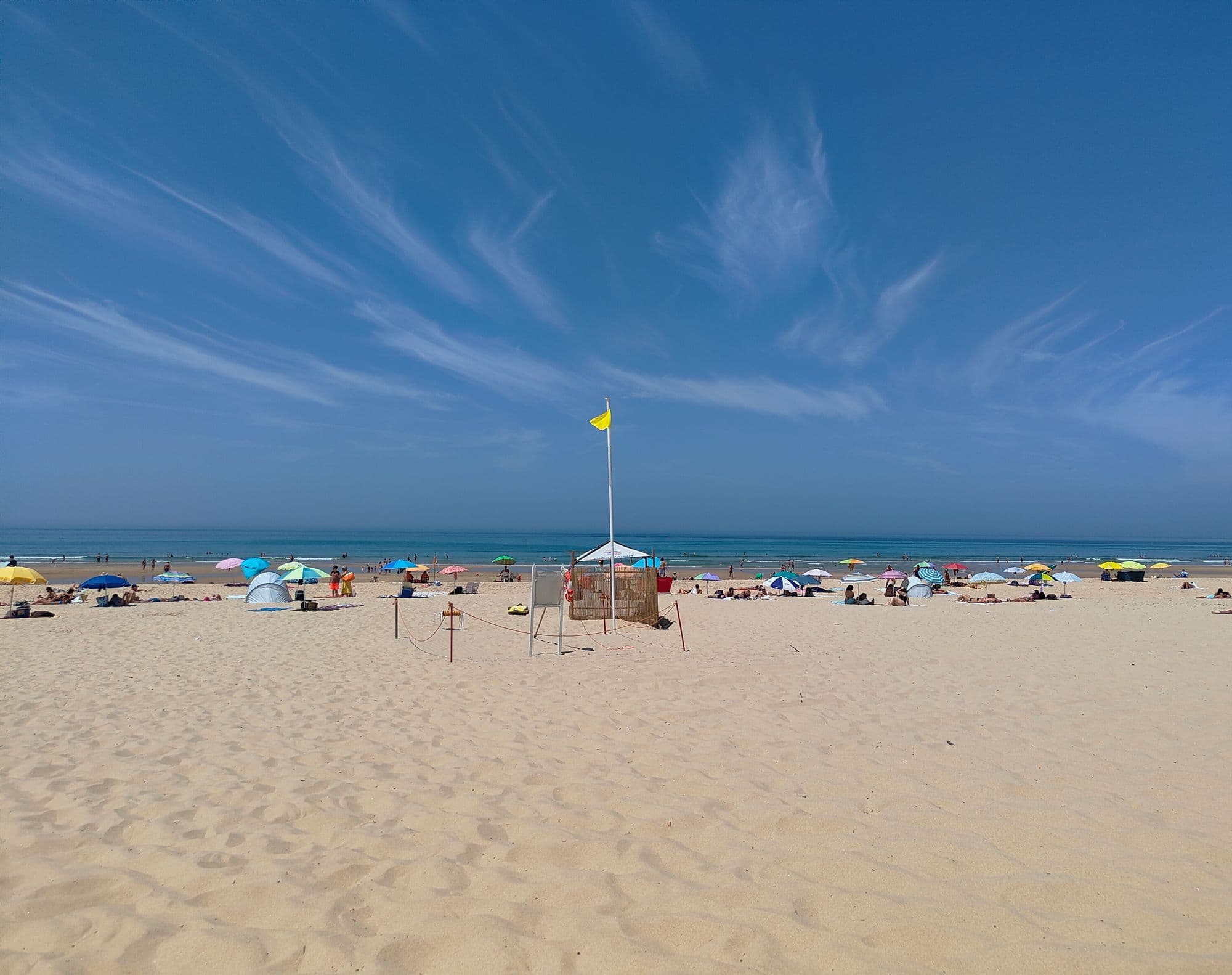 A wide sandy beach at Costa da Caparica under a bright blue sky, with sun umbrellas and a yellow lifeguard flag.