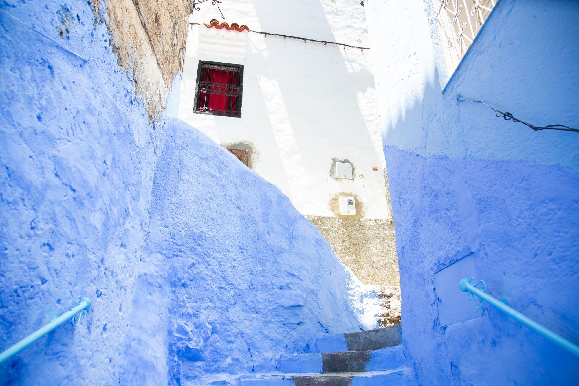 A narrow stepped lane between walls painted bright blue in Chefchaouen, with a red window frame above.