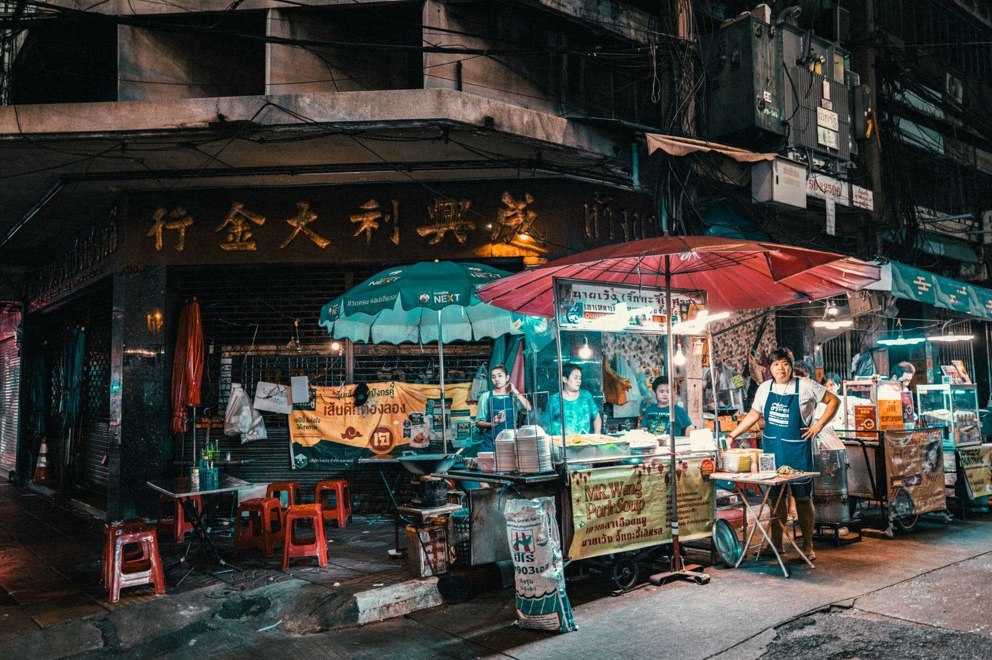 A street-food cart under red and green umbrellas on a Bangkok corner at night, with cooks working under bare bulbs.