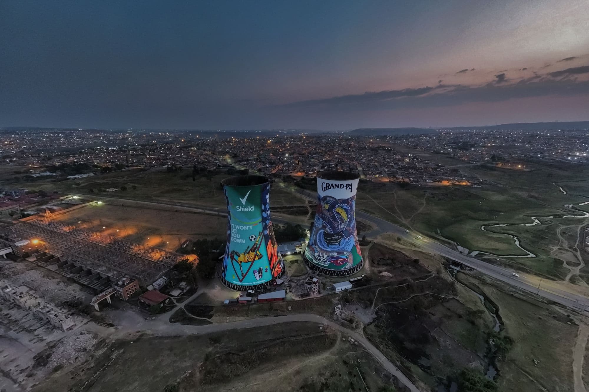 Aerial view of the two painted Orlando cooling towers in Soweto at twilight, with lights spreading across the township behind.
