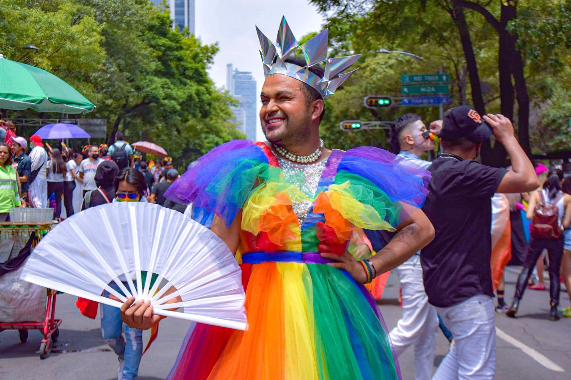 A smiling marcher in a rainbow tulle dress and silver crown holds a white fan on a tree-lined Mexico City avenue.