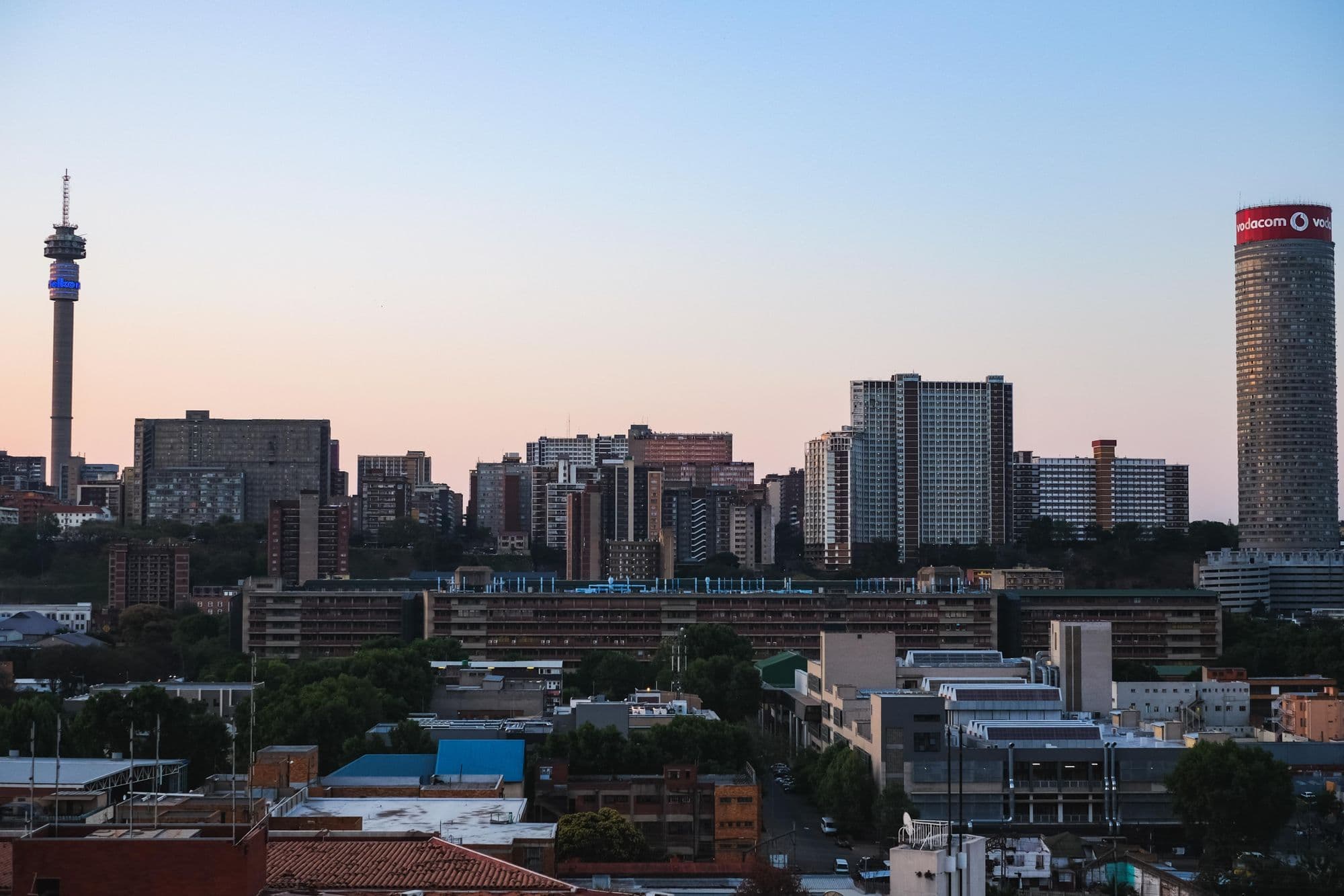 The Johannesburg skyline at dusk under a pink sky, with the Hillbrow tower and Ponte Tower rising above lower buildings.