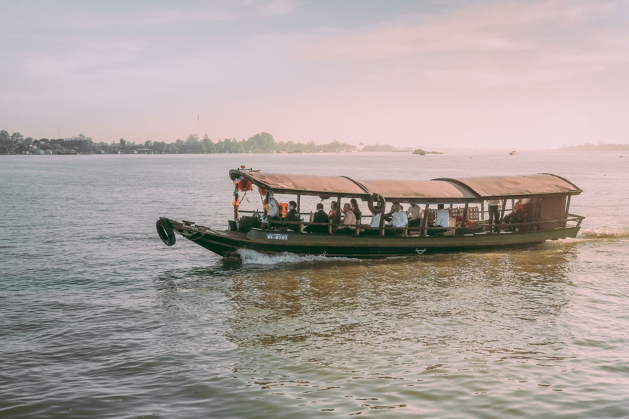 A covered wooden boat carrying passengers across the wide Mekong River in Vĩnh Long at sunset.