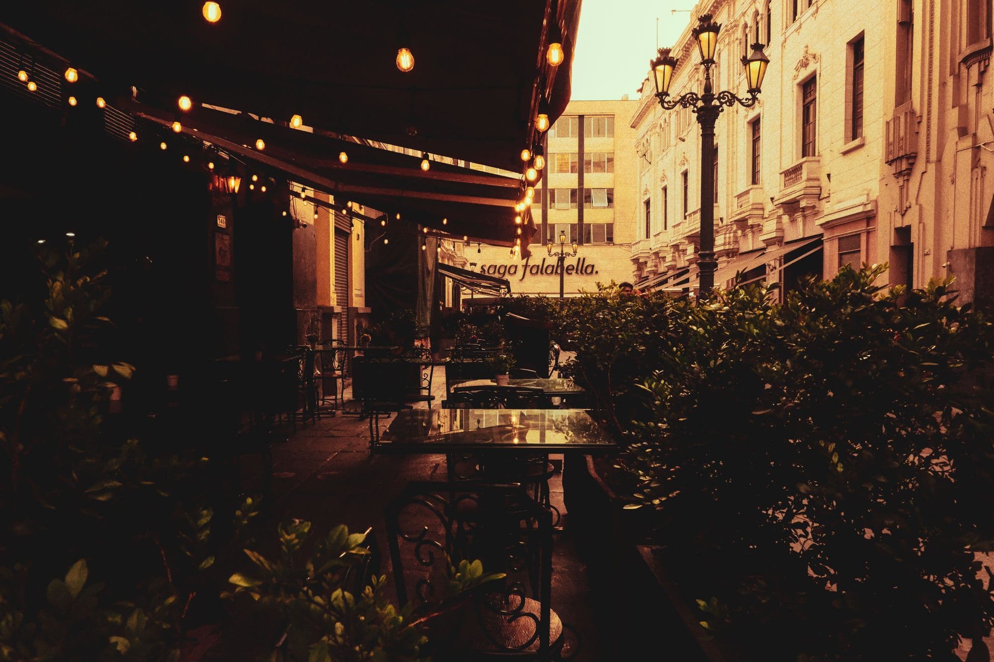 Tables sit outside a warmly lit café on a city street at dusk.
