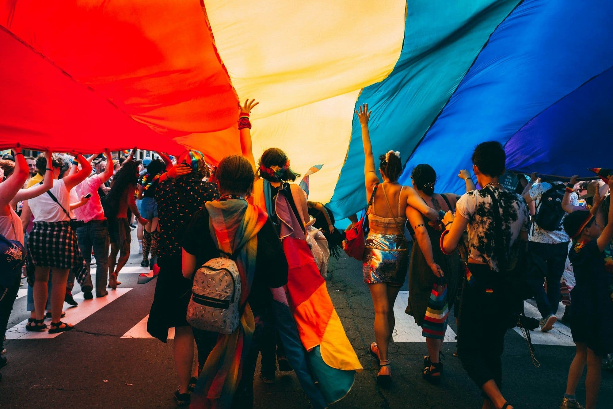 Marchers walk beneath a wide rainbow flag on a city street during Pride.