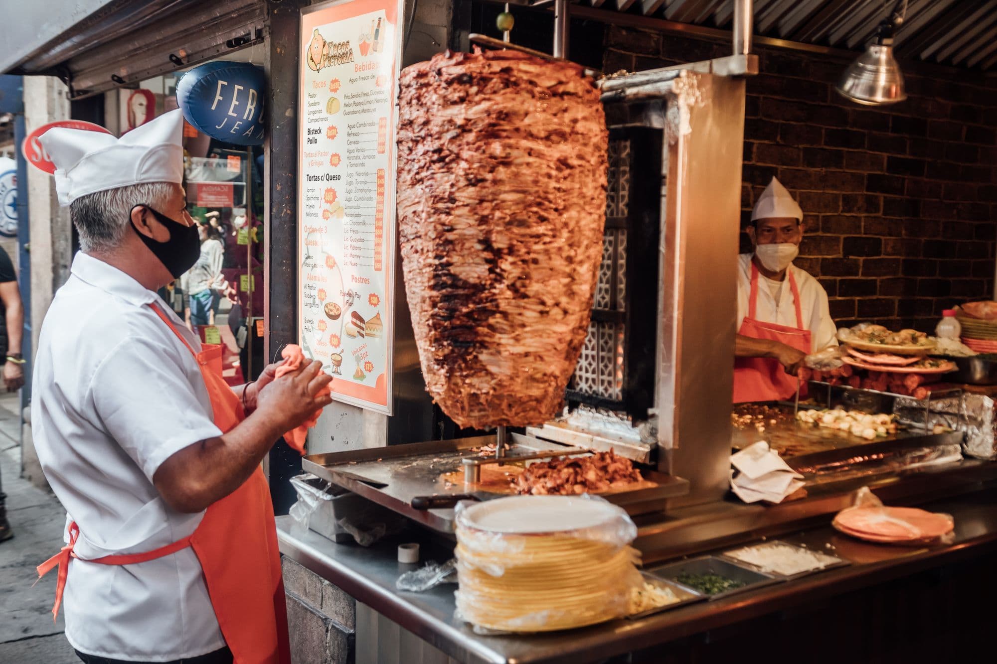 A taquero in an orange apron beside a tall spit of al pastor pork, with a stack of tortillas on the counter.