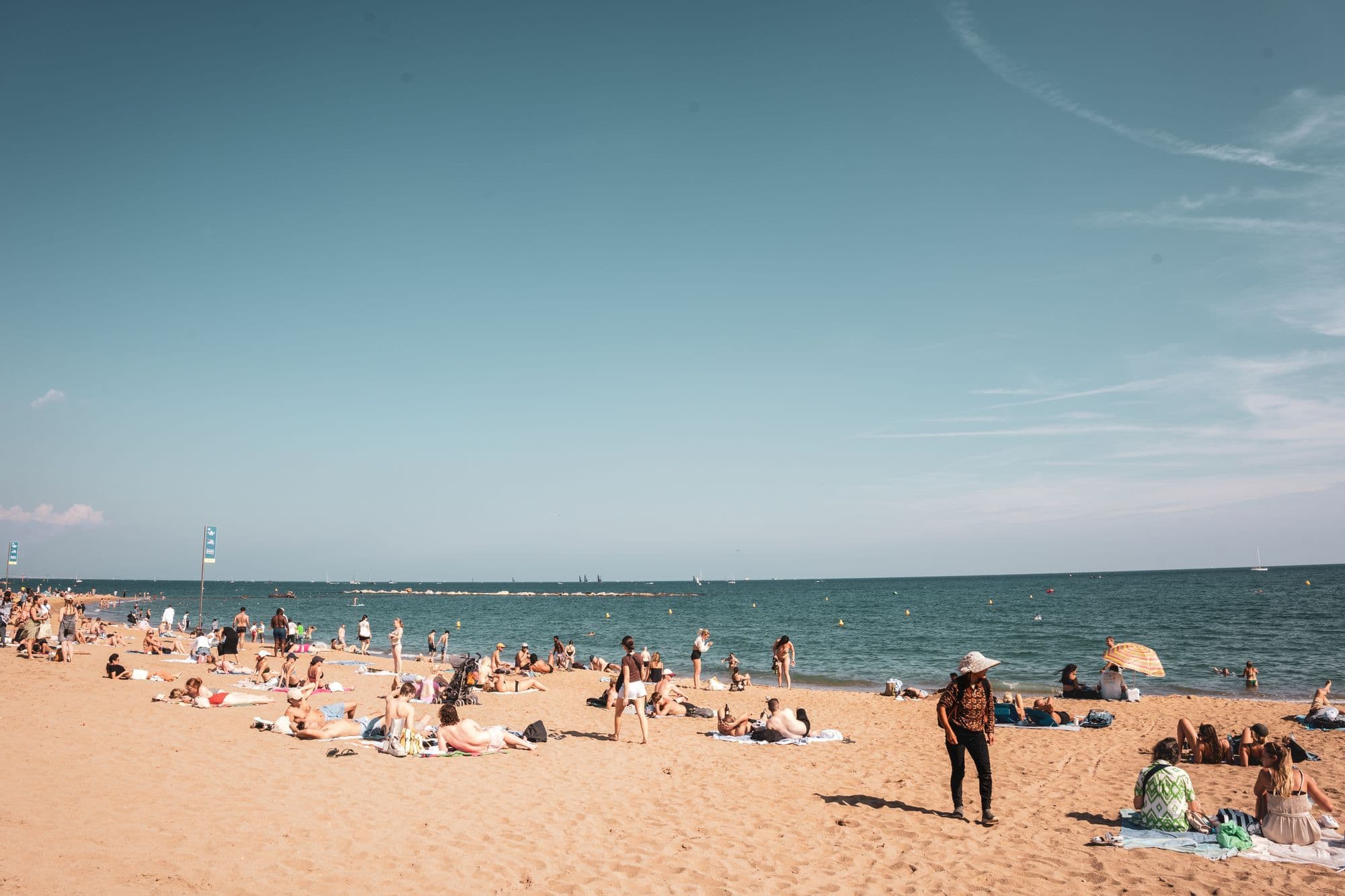 Sunbathers and swimmers fill a broad sandy Barcelona beach under a clear blue sky.