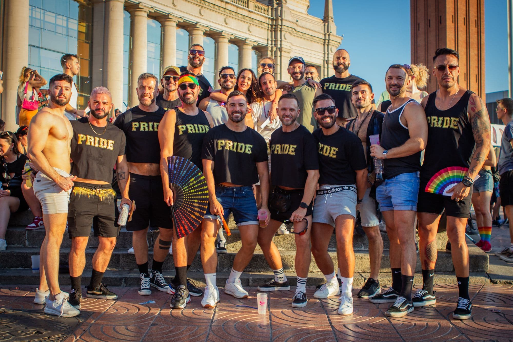 A group of men in matching black Pride T-shirts posing together on a Barcelona street on a sunny day.