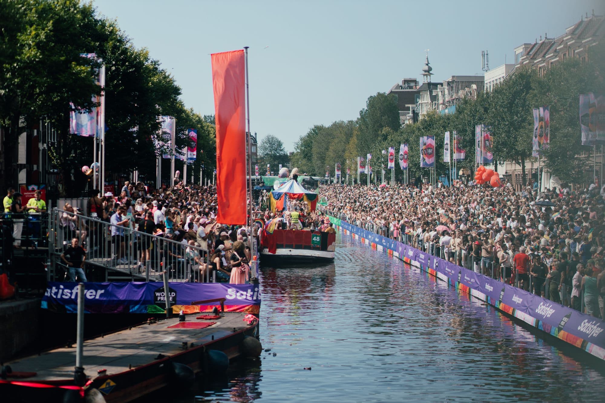 A dense Pride crowd watches decorated boats pass along an Amsterdam canal.