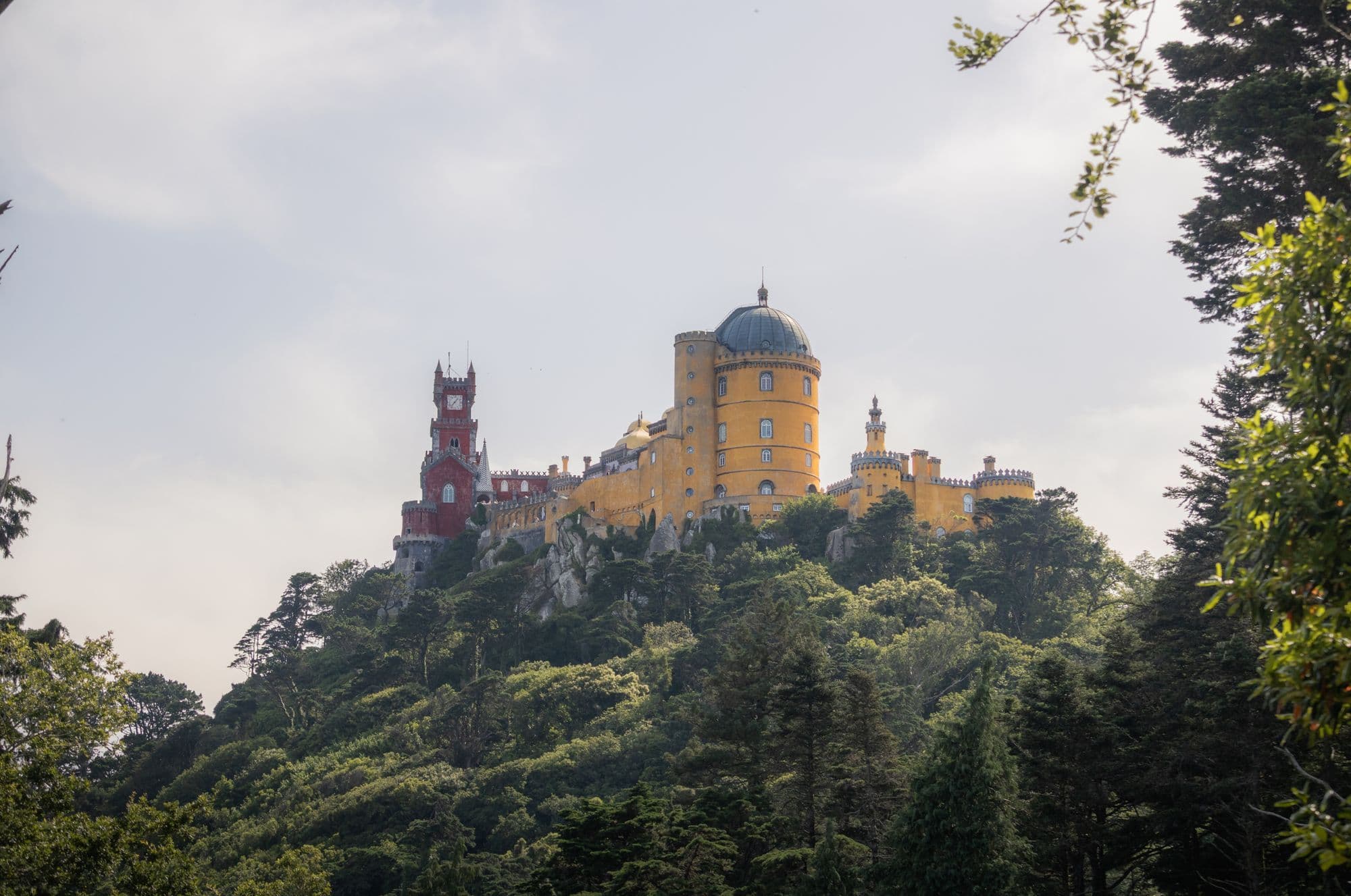 The yellow and red towers of Pena Palace rise above a forested hilltop under a pale sky.