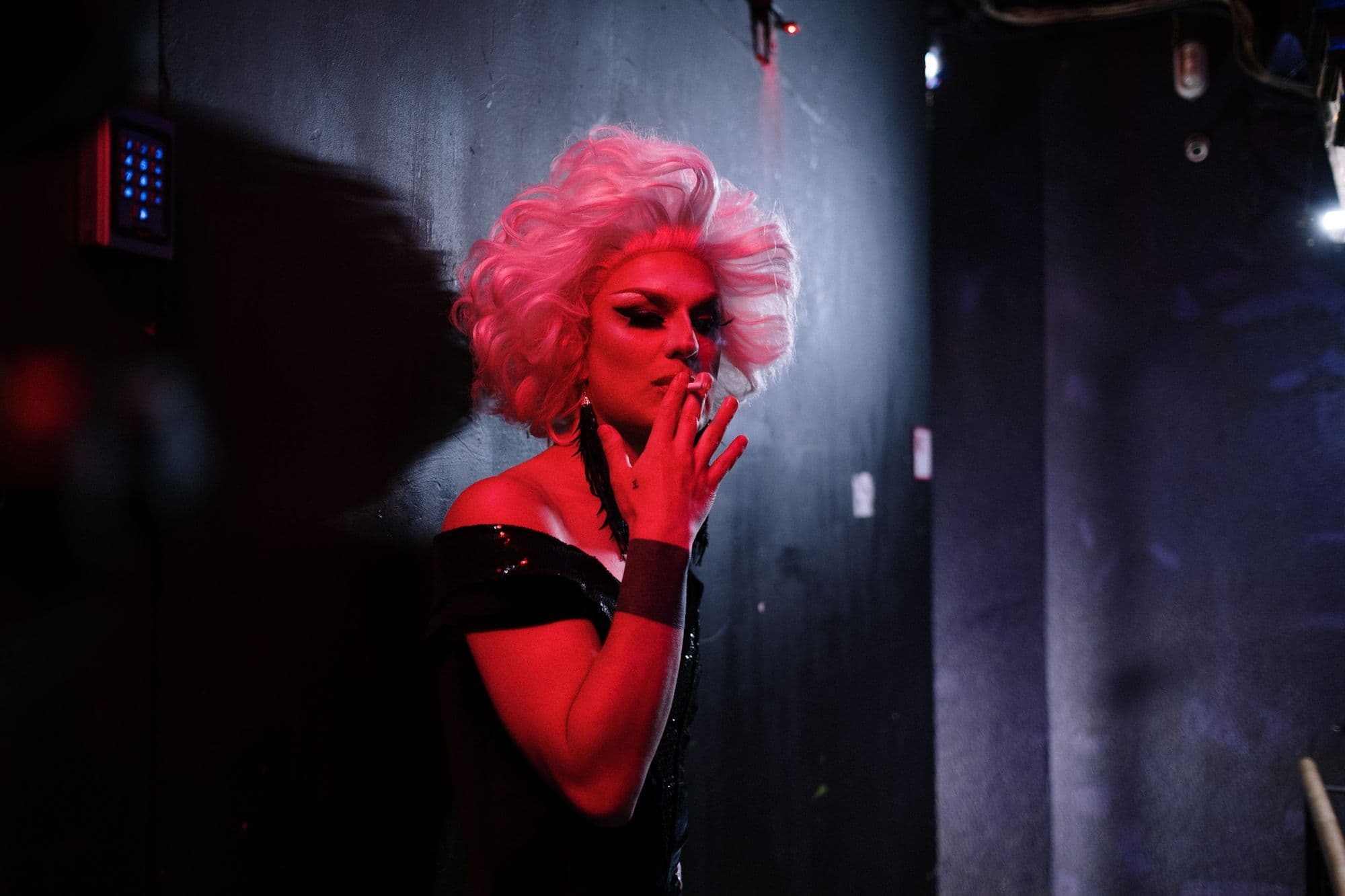 A drag performer in a white wig and black dress standing under red and blue club lights.