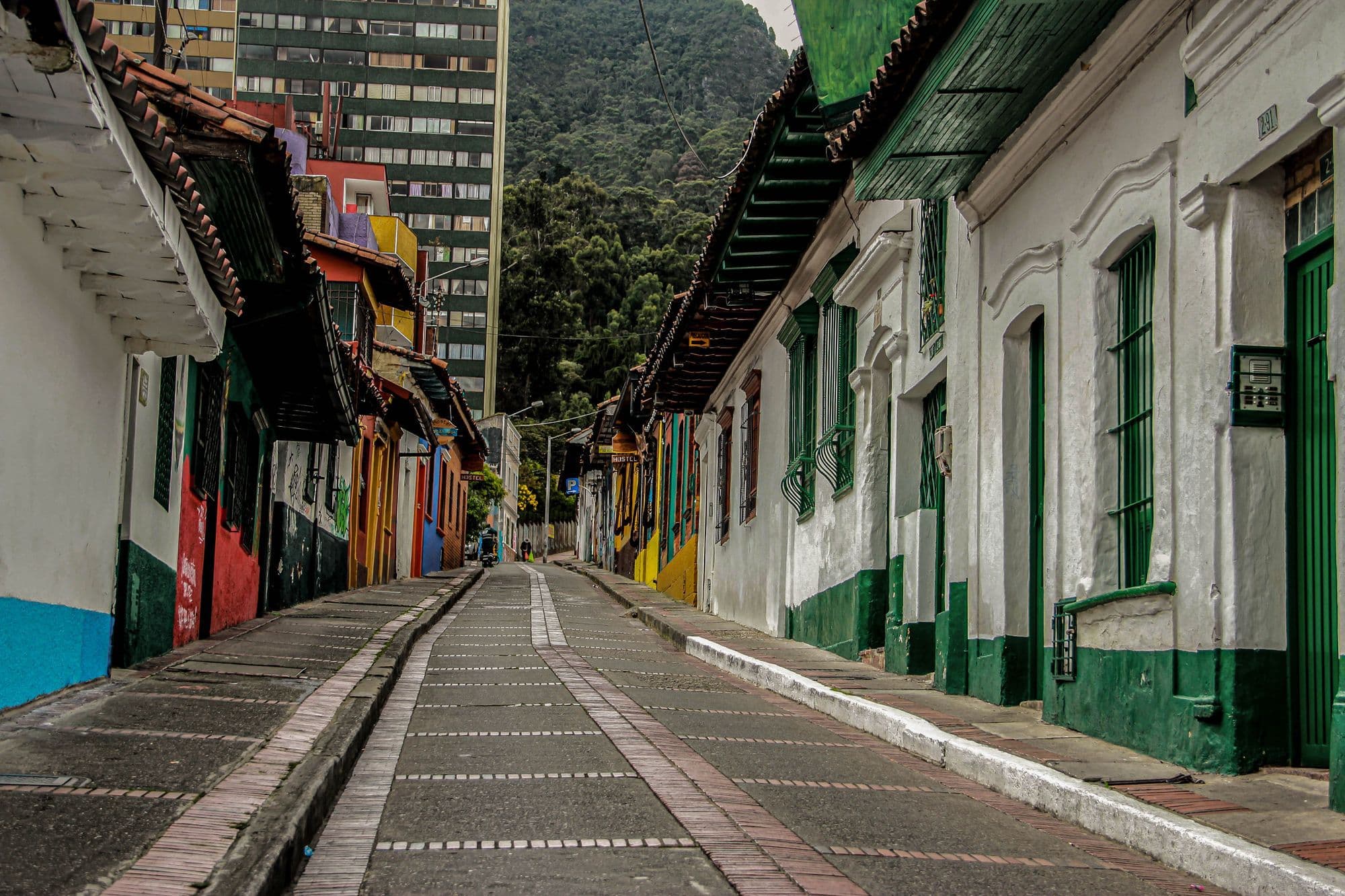 A sloping cobbled street in La Candelaria, Bogotá, lined with colourful colonial houses with green balconies and wooden doors.