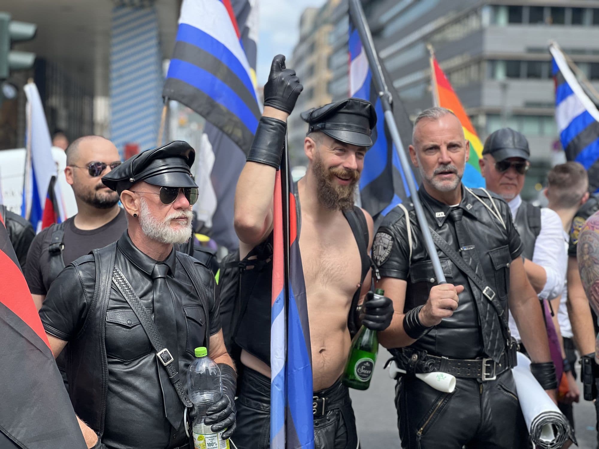 Bearded men in black leather caps, harnesses and gloves holding blue-and-black leather Pride flags during Berlin's Pride parade.