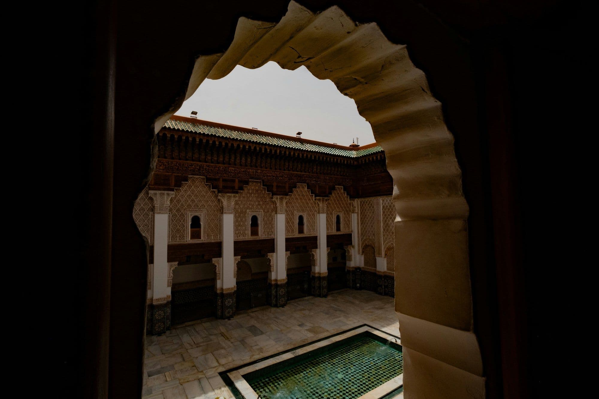 A courtyard with carved arcades and a long rectangular pool, seen through a scalloped archway in Marrakech.