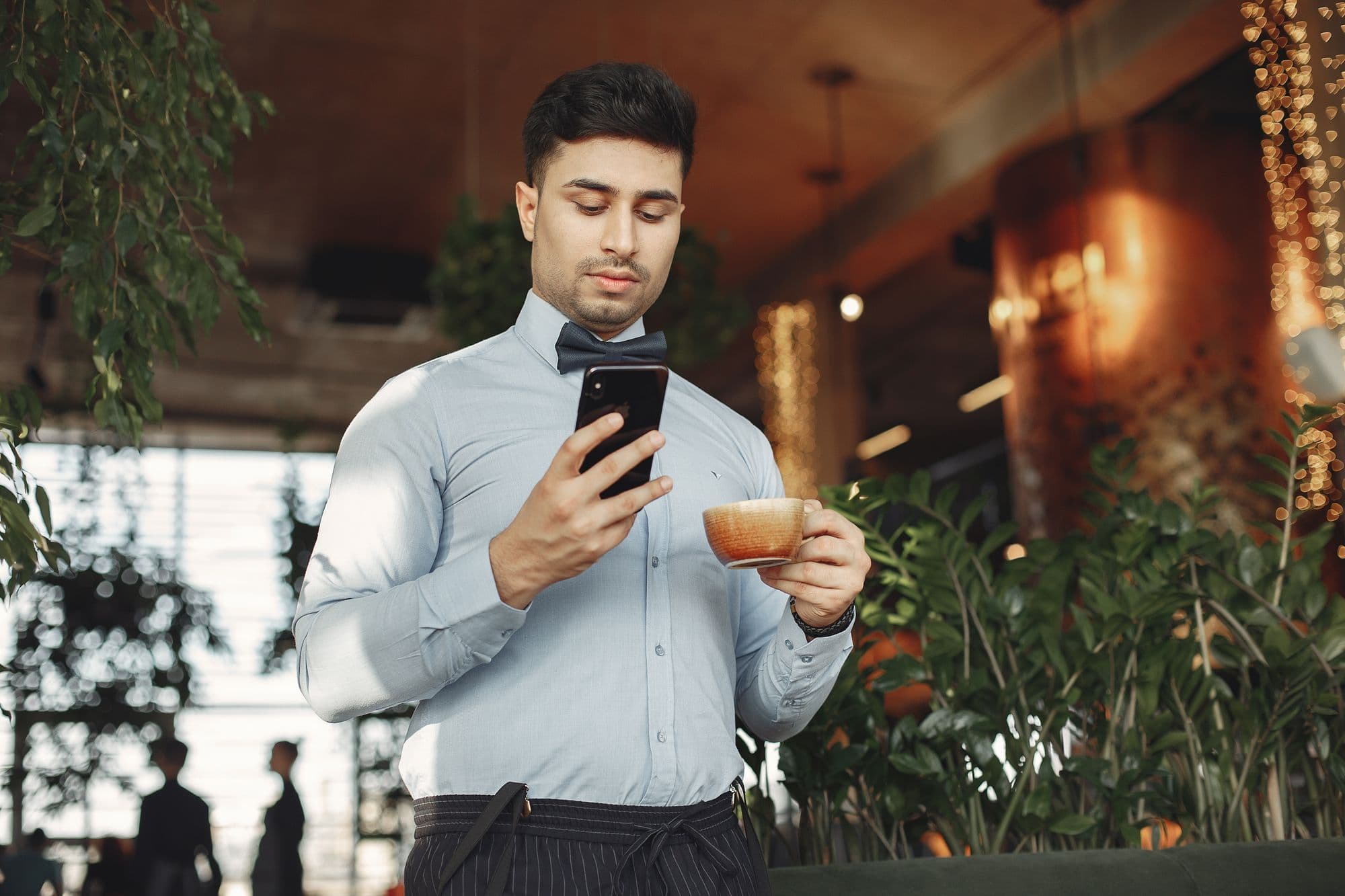 A young man checks his smartphone at a café table with a coffee in front of him.