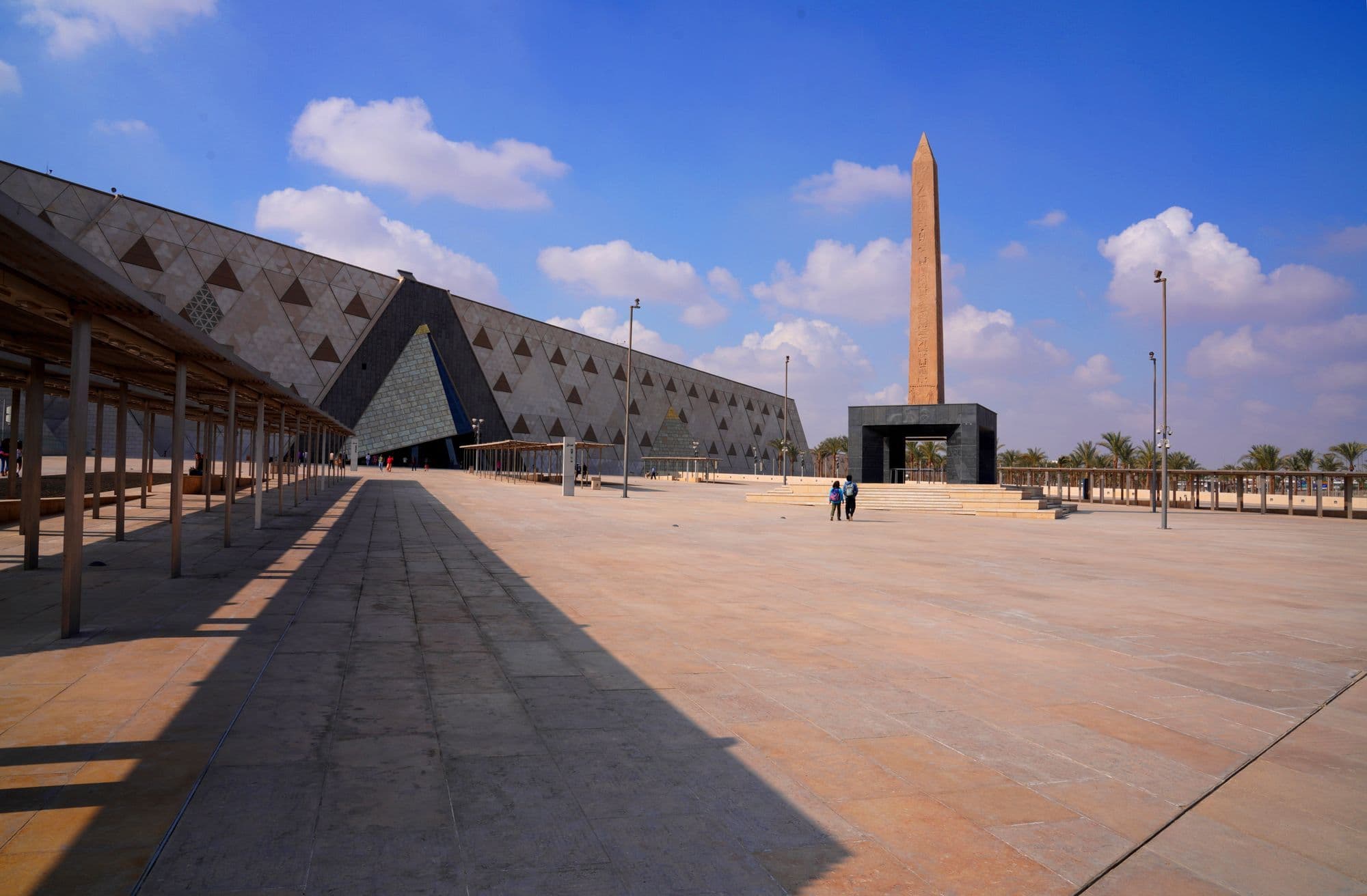 The angled glass-and-stone facade of the Grand Egyptian Museum with a tall obelisk standing in the paved plaza in front.