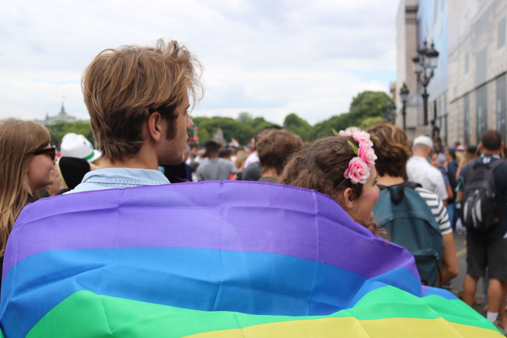 Paris Pride participants wrapped in a rainbow flag move through a crowded city street.