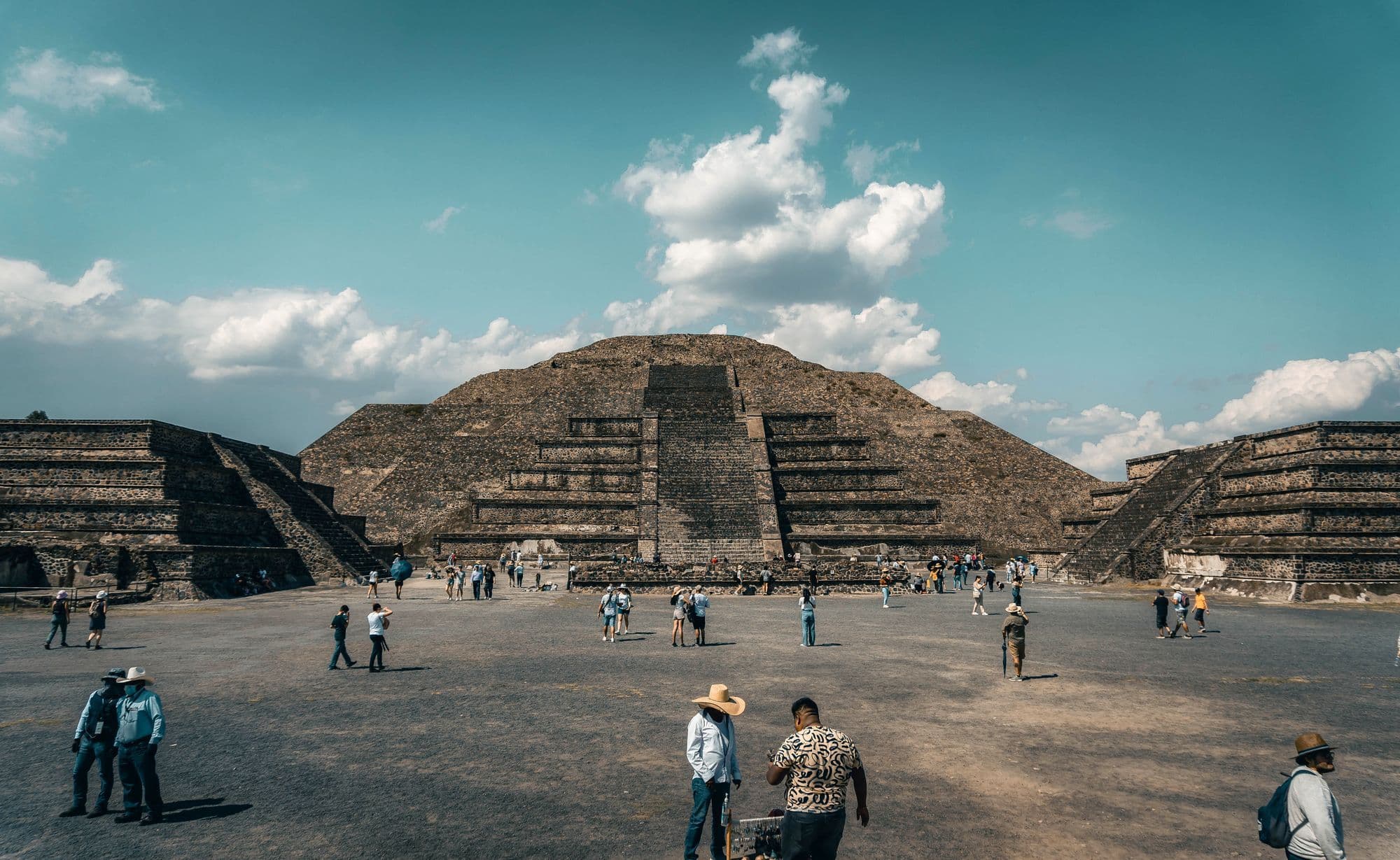 The Pyramid of the Moon at Teotihuacan under a blue sky, visitors in hats crossing the dusty plaza in front.