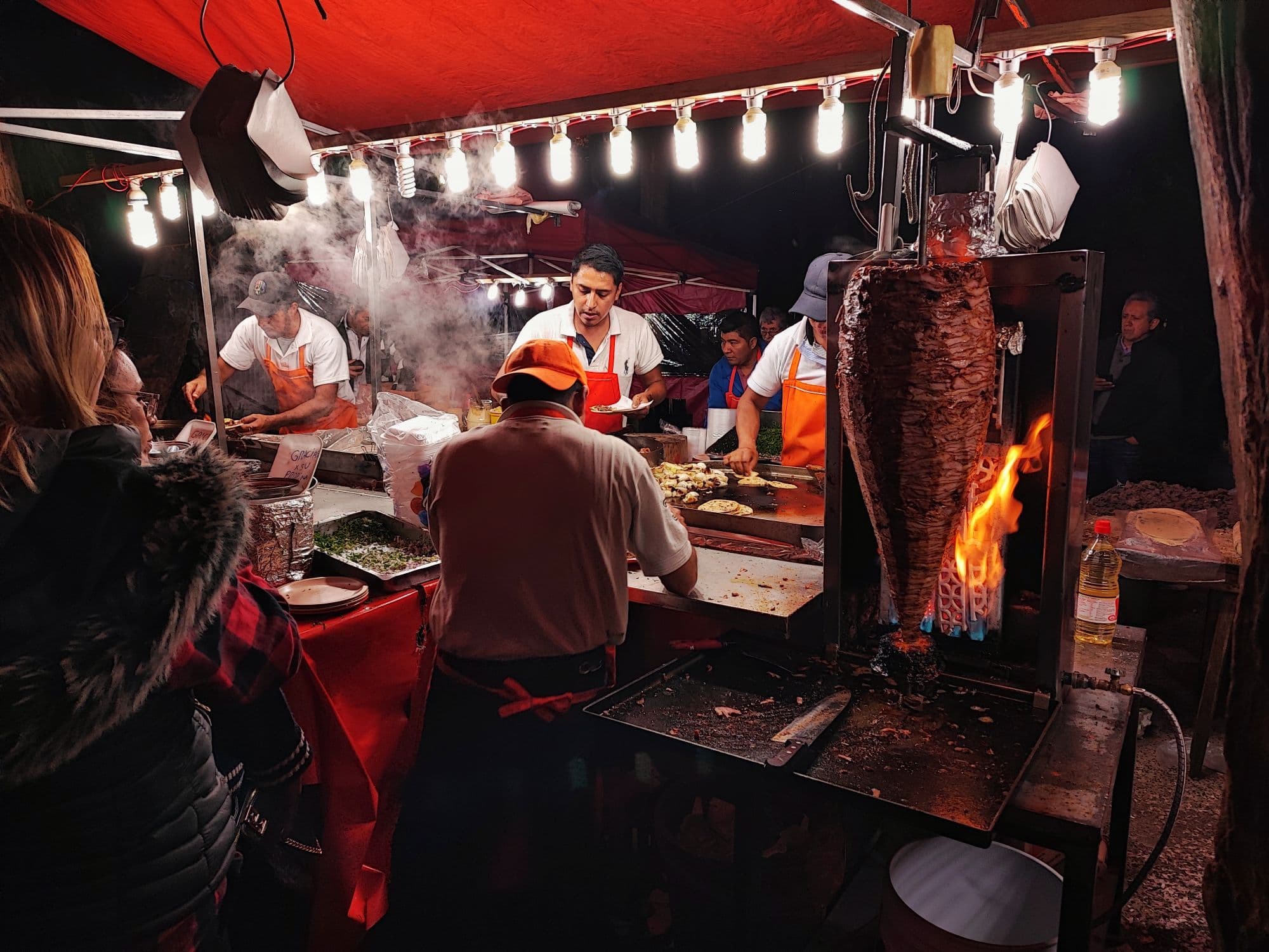 Cooks in orange aprons at a night taco stall under string lights, a spit of al pastor pork beside a flame.