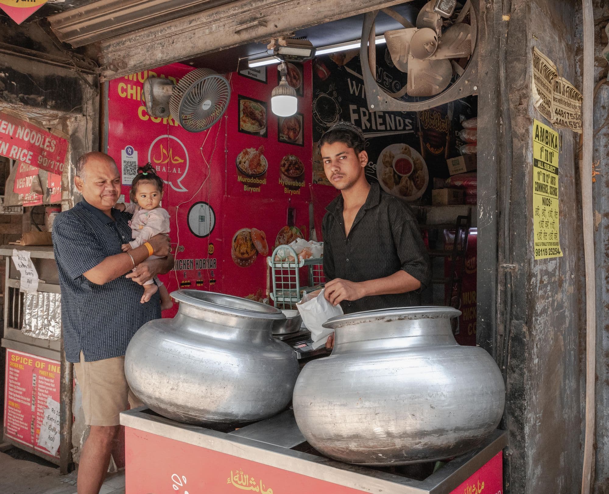 A smiling vendor serves from two large polished metal pots at a red-painted market stall in Delhi.