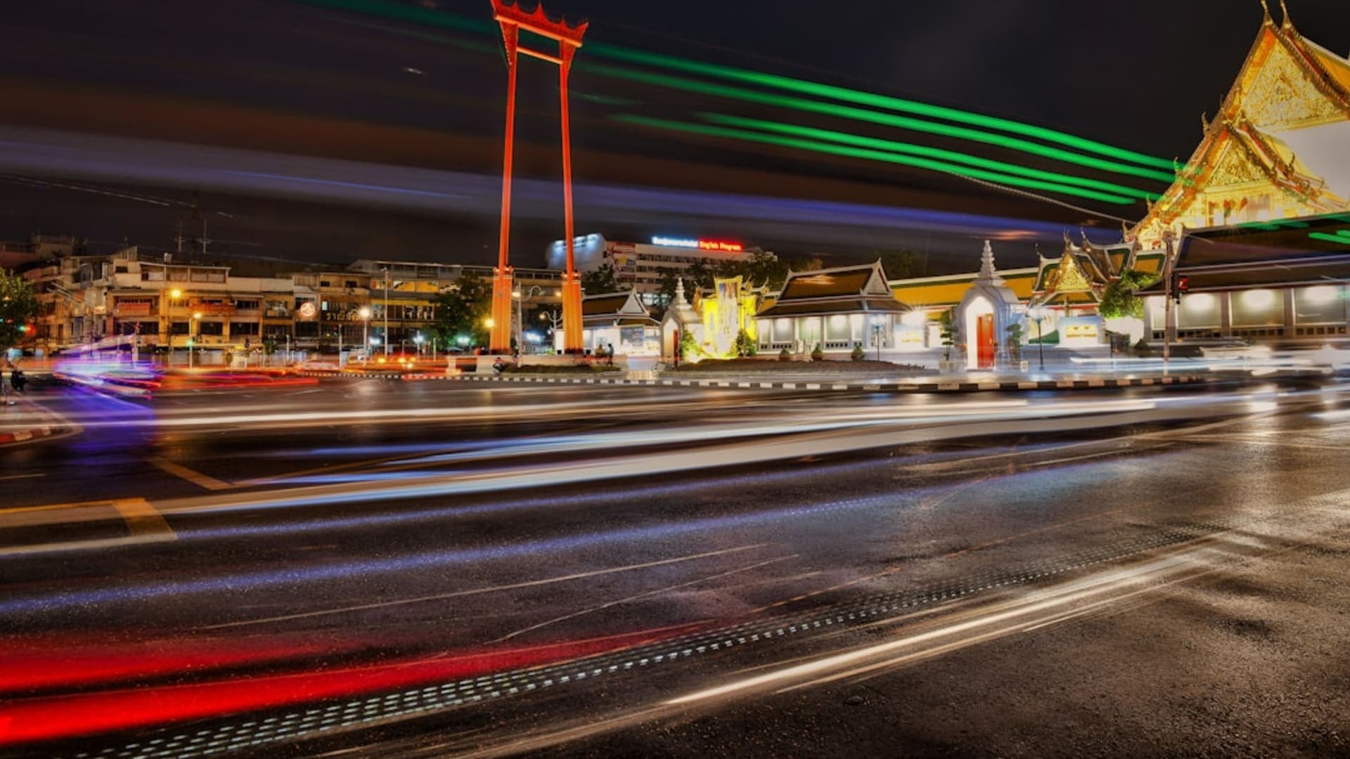 A long-exposure night shot of Bangkok traffic light trails streaming past an illuminated temple gate.