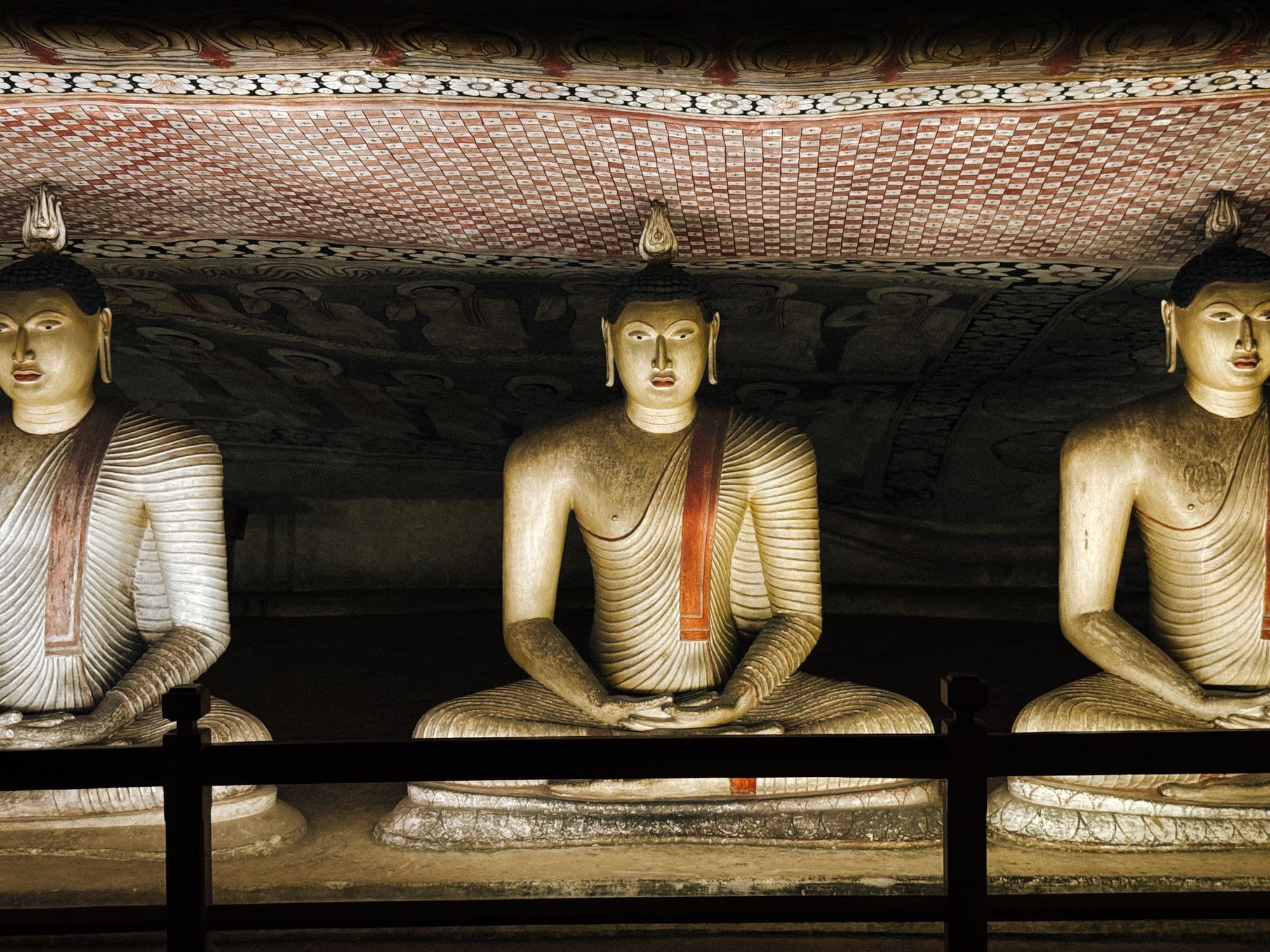 A row of large seated Buddha statues with painted robes beneath a patterned rock ceiling inside the Dambulla cave temple.