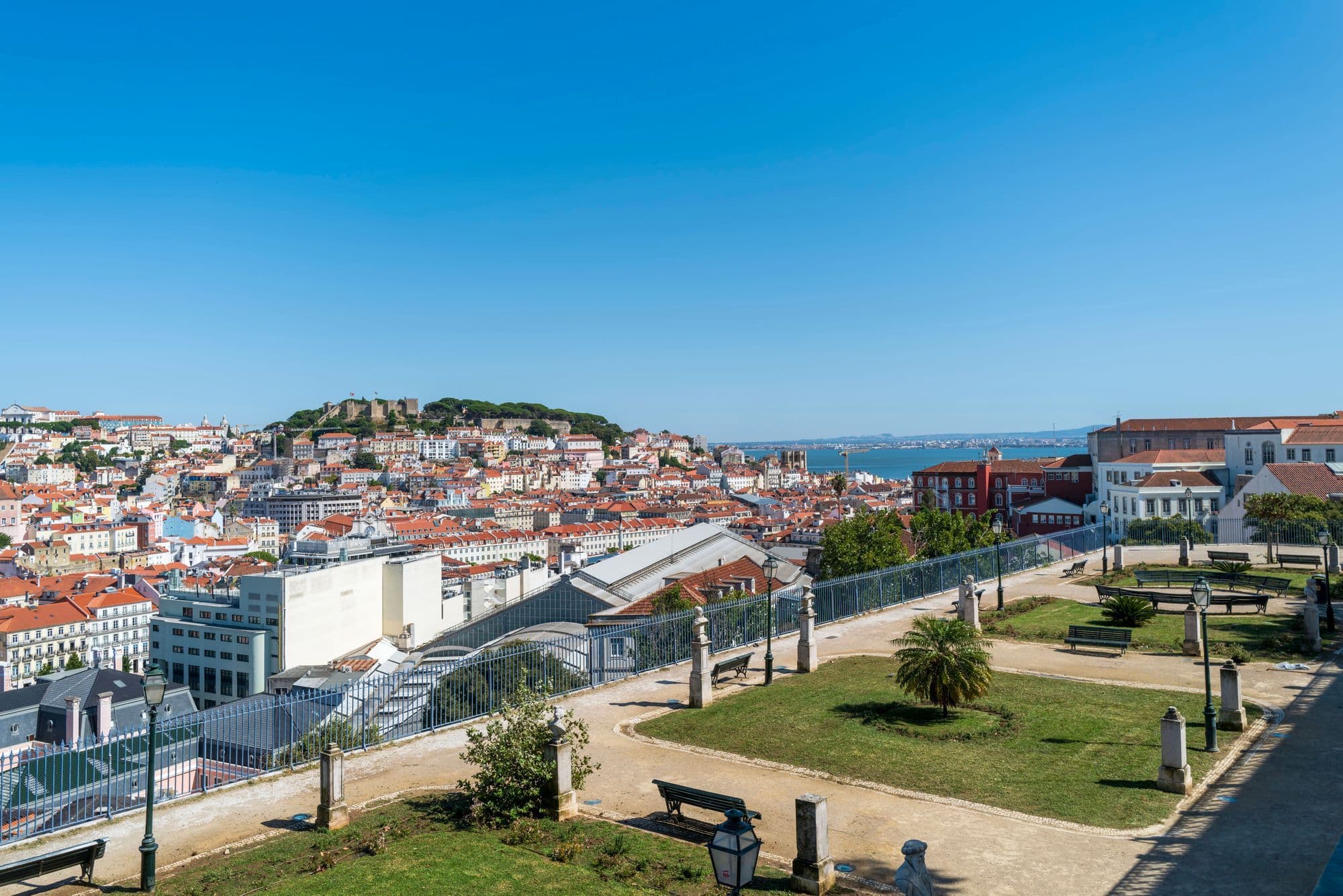 A terraced garden viewpoint with stone busts and a palm looks across red rooftops to São Jorge Castle on the opposite hill.