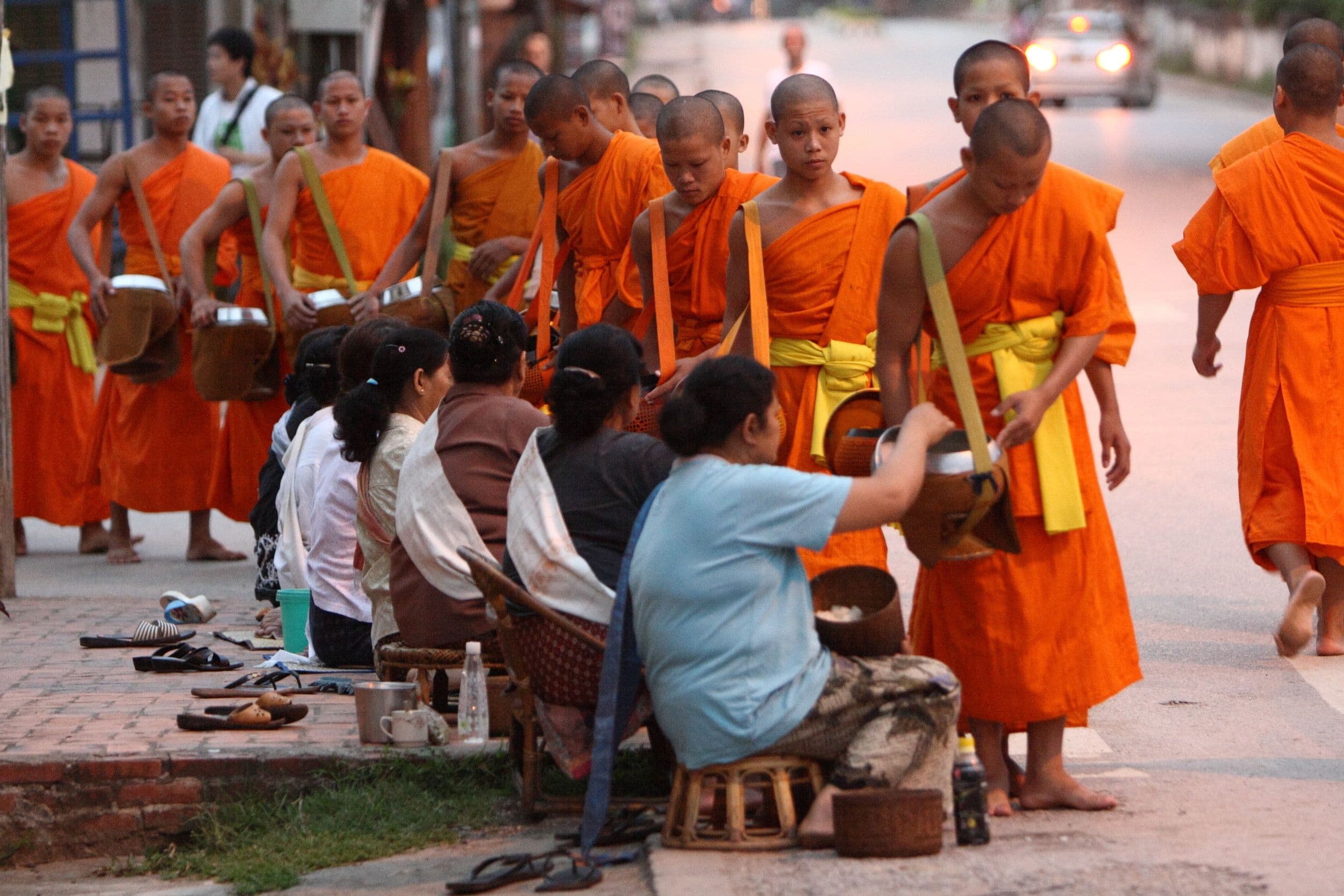 A line of monks in orange robes walking past people kneeling on low stools to give alms at dawn in Luang Prabang.