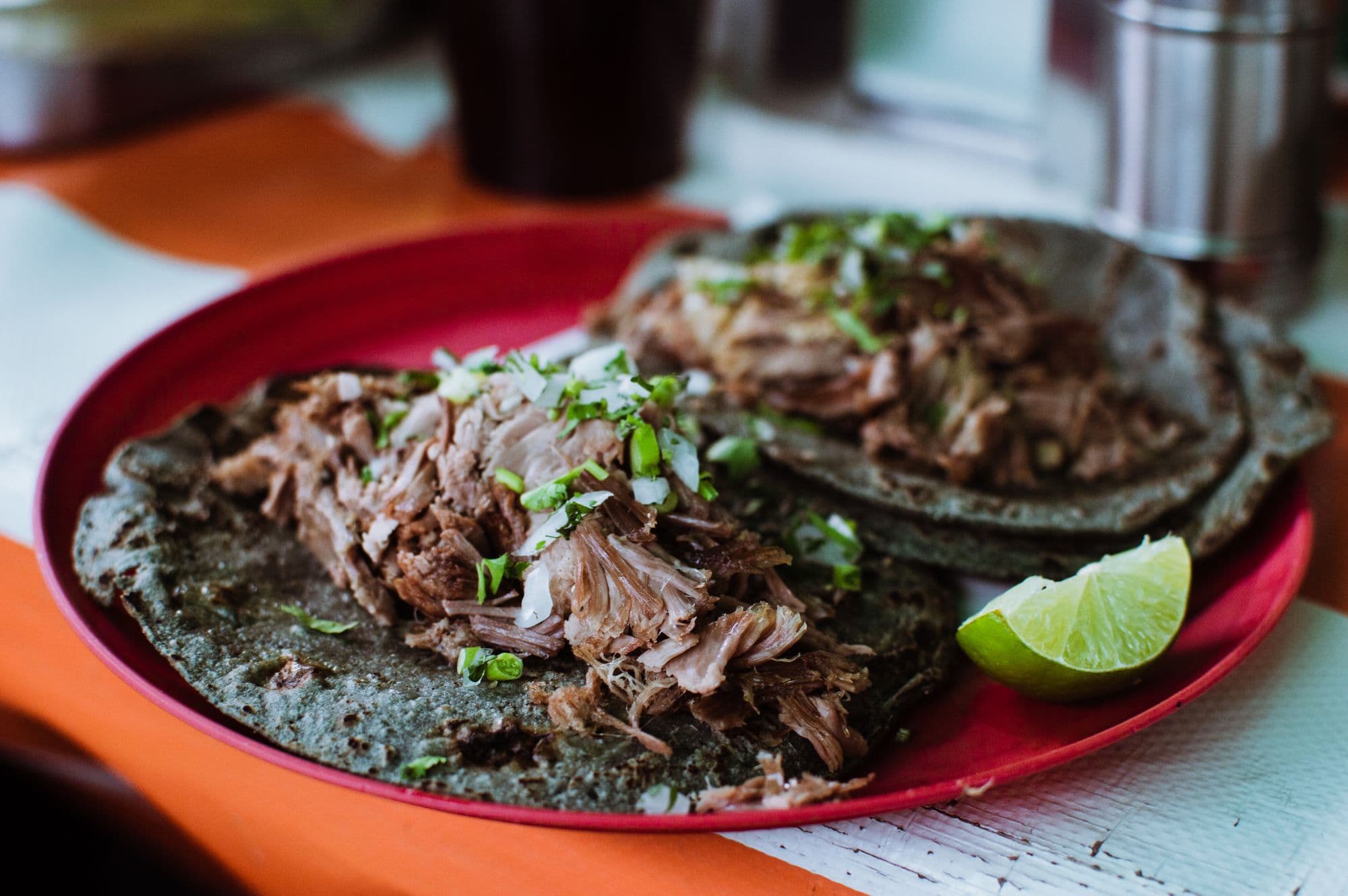Two barbacoa tacos on blue-corn tortillas with chopped onion and coriander, served on a red plate with a lime wedge.