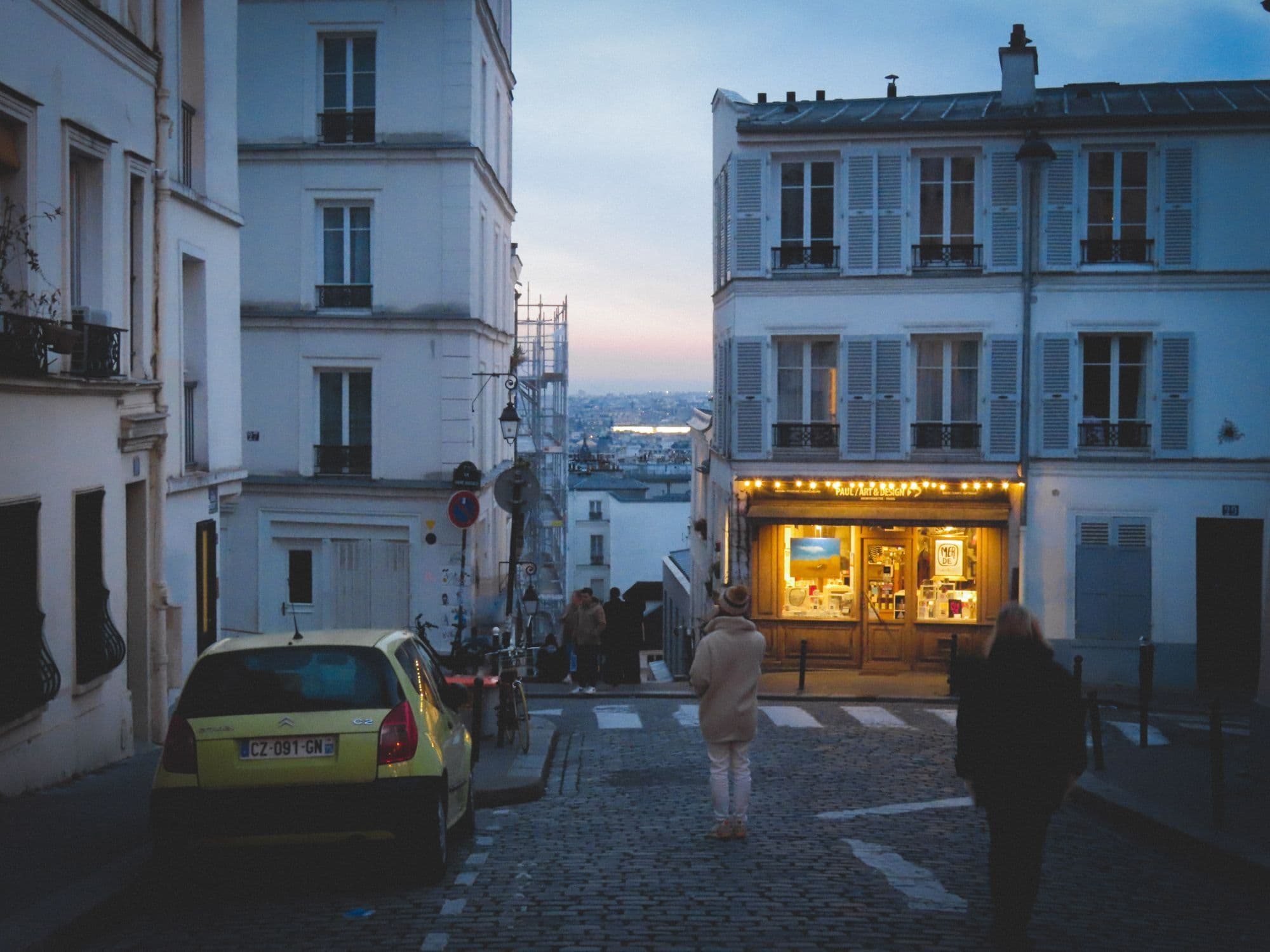 People walk past glowing shopfronts on a narrow Montmartre street after dark.