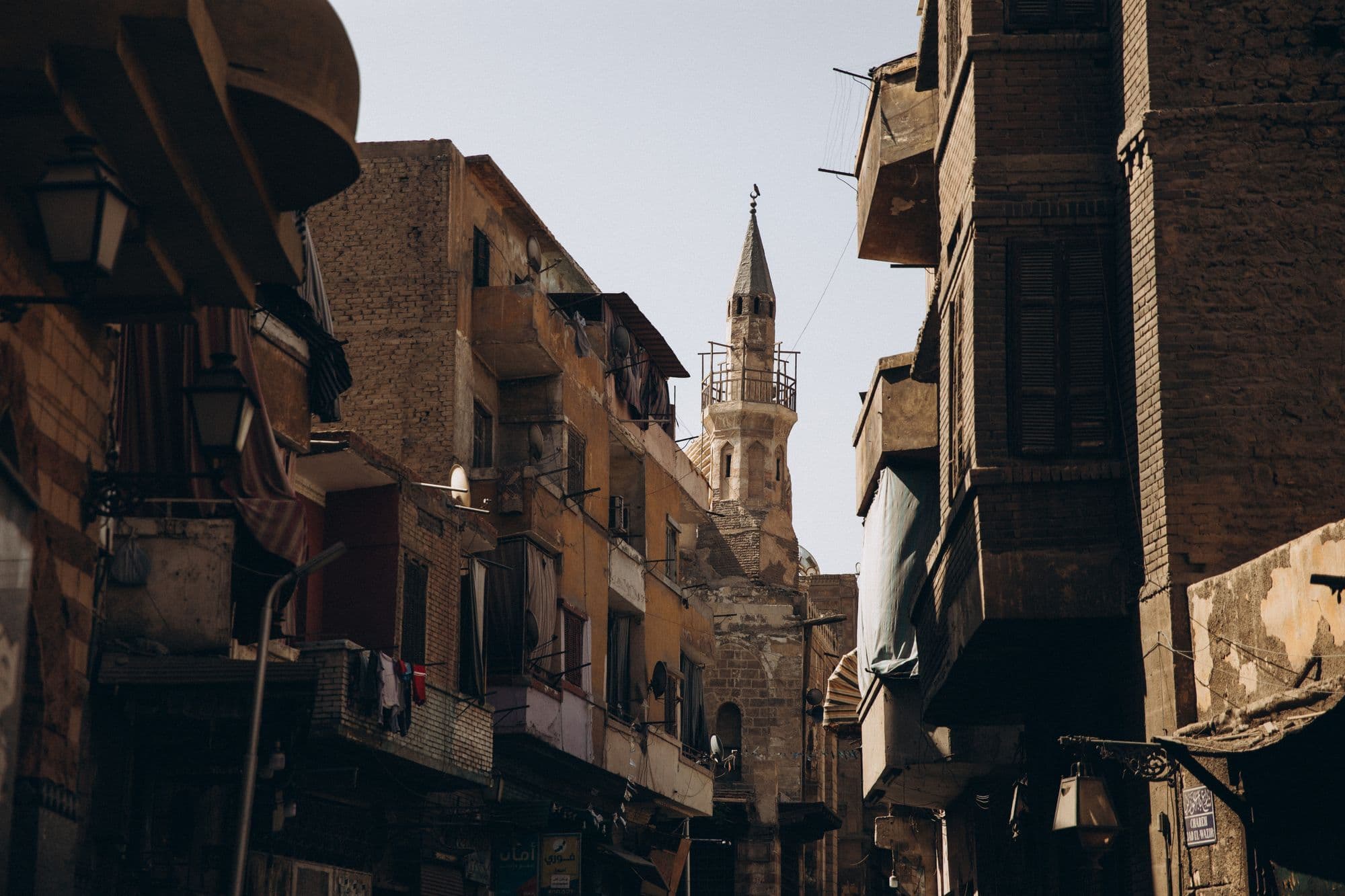 A narrow street in historic Cairo lined with tall weathered buildings, with a minaret rising at the far end.