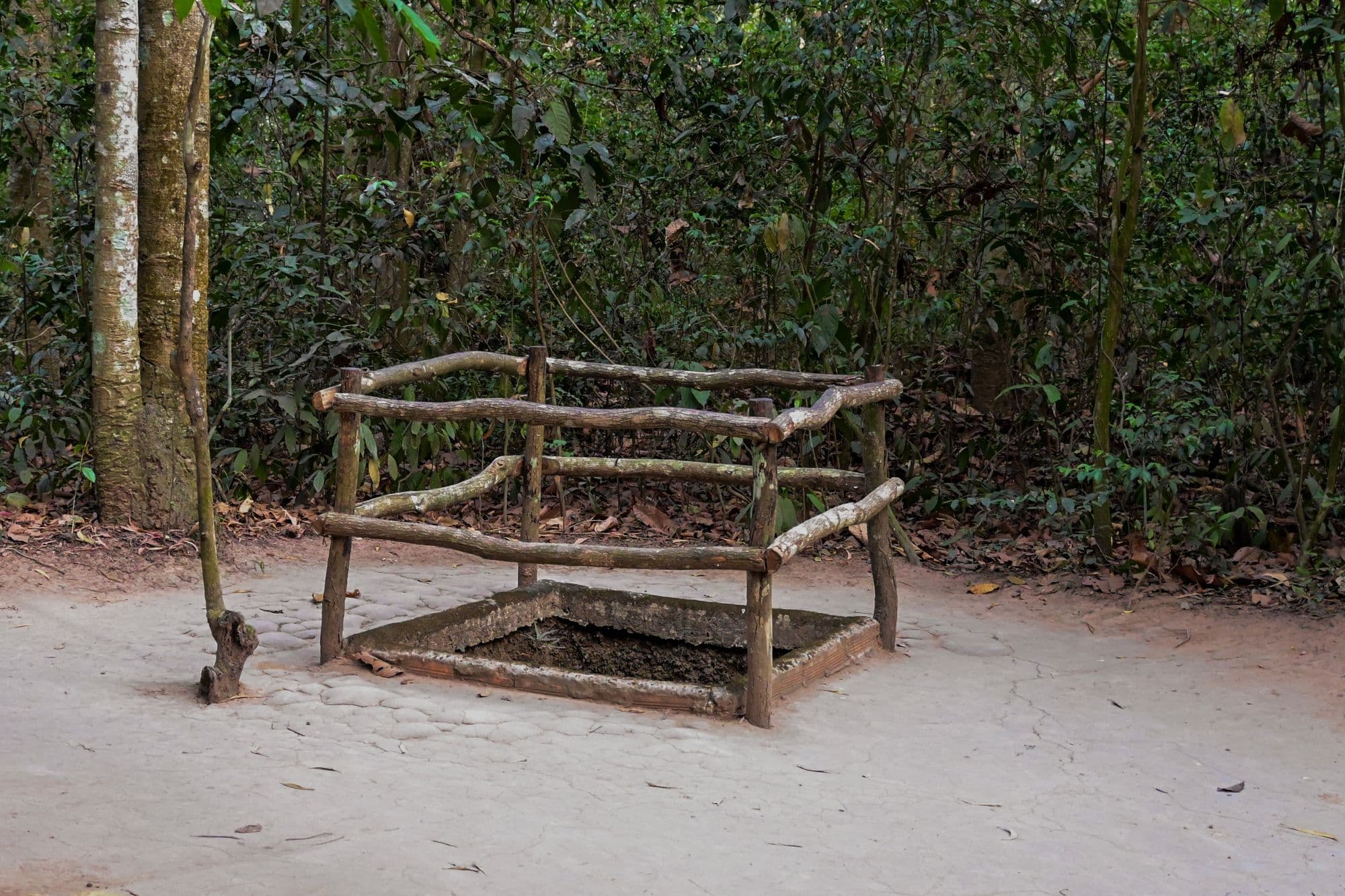 A square tunnel opening in the forest floor framed by a low railing of branches at the Cu Chi Tunnels.