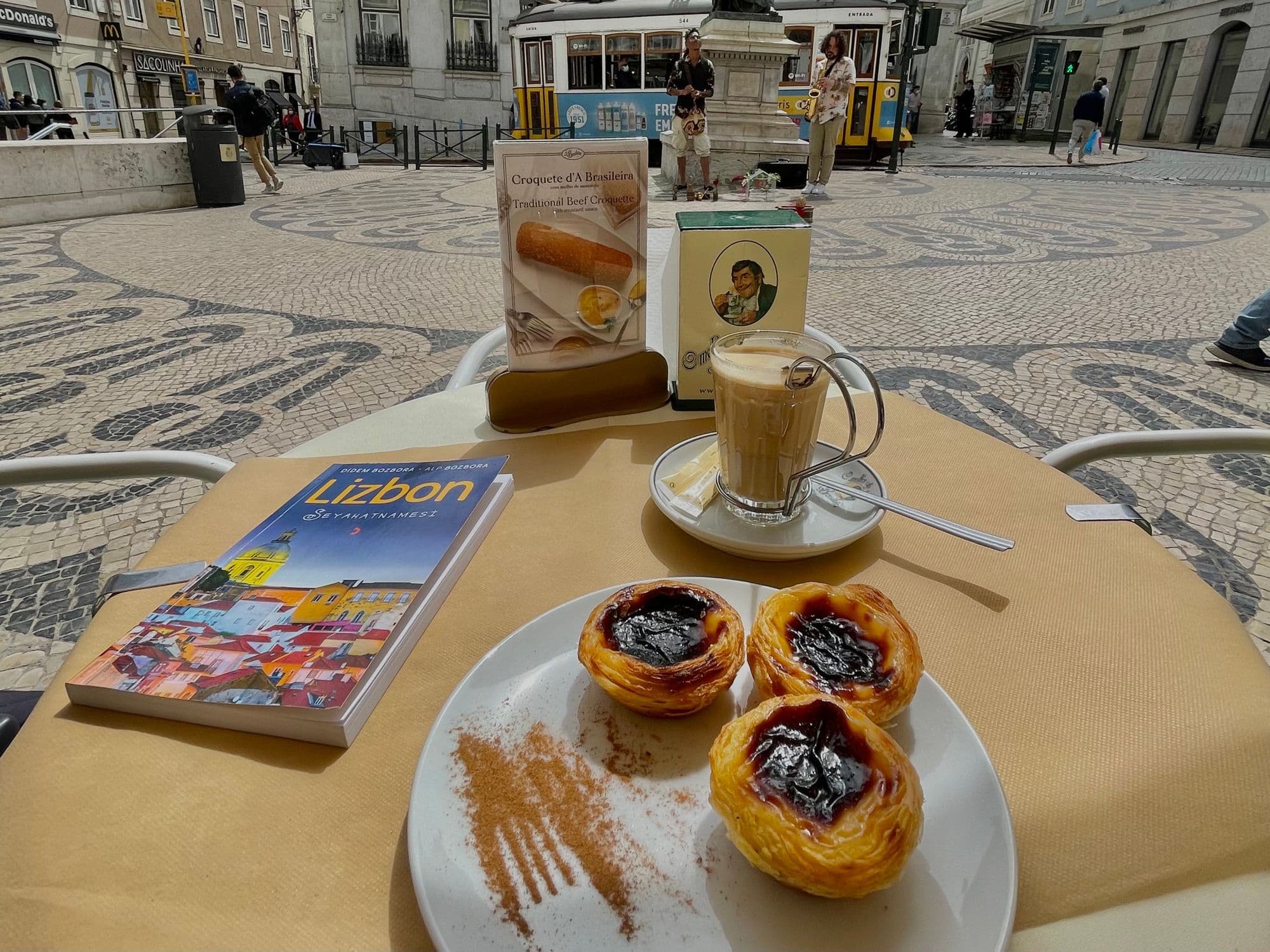 Three custard tarts dusted with cinnamon beside a milky coffee on a café table, with a yellow tram parked behind.