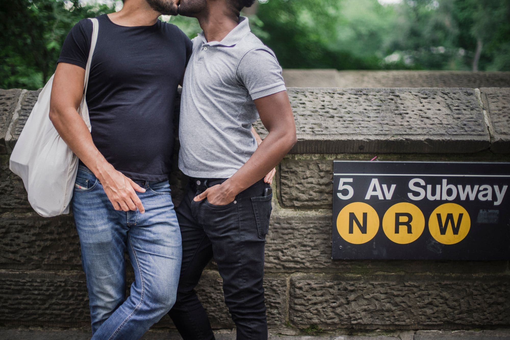 Two men kissing beside a subway entrance on a sunny New York street.
