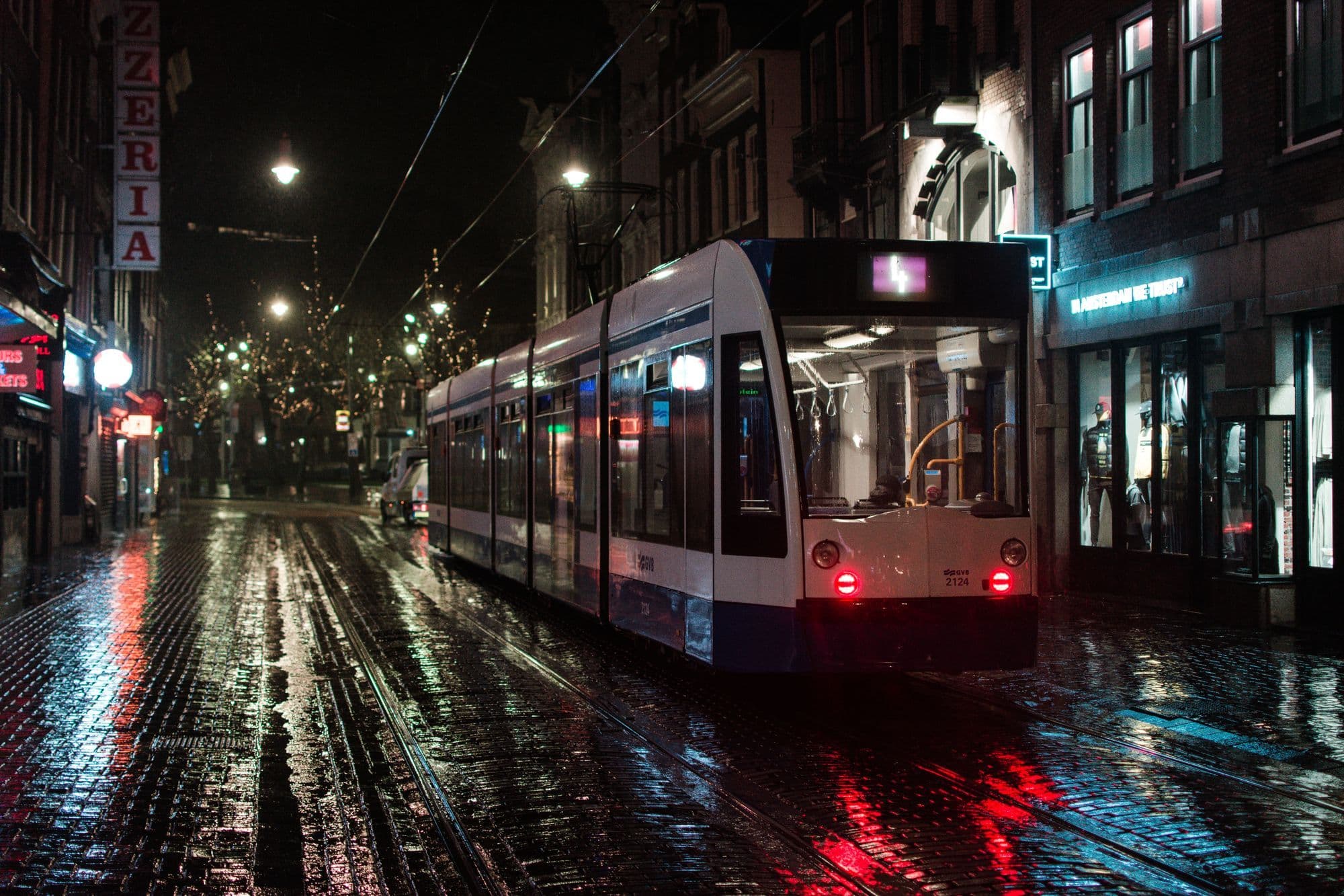 A white and blue tram on a wet, lamp-lit shopping street in Amsterdam at night, rails reflecting red tail lights.