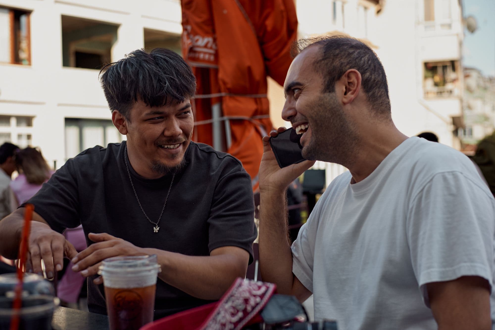 Two men smiling and talking over drinks at an outdoor café table.
