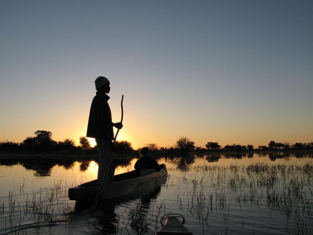 The silhouette of a poler standing in a mokoro dugout canoe on still water in the Okavango Delta at sunset.