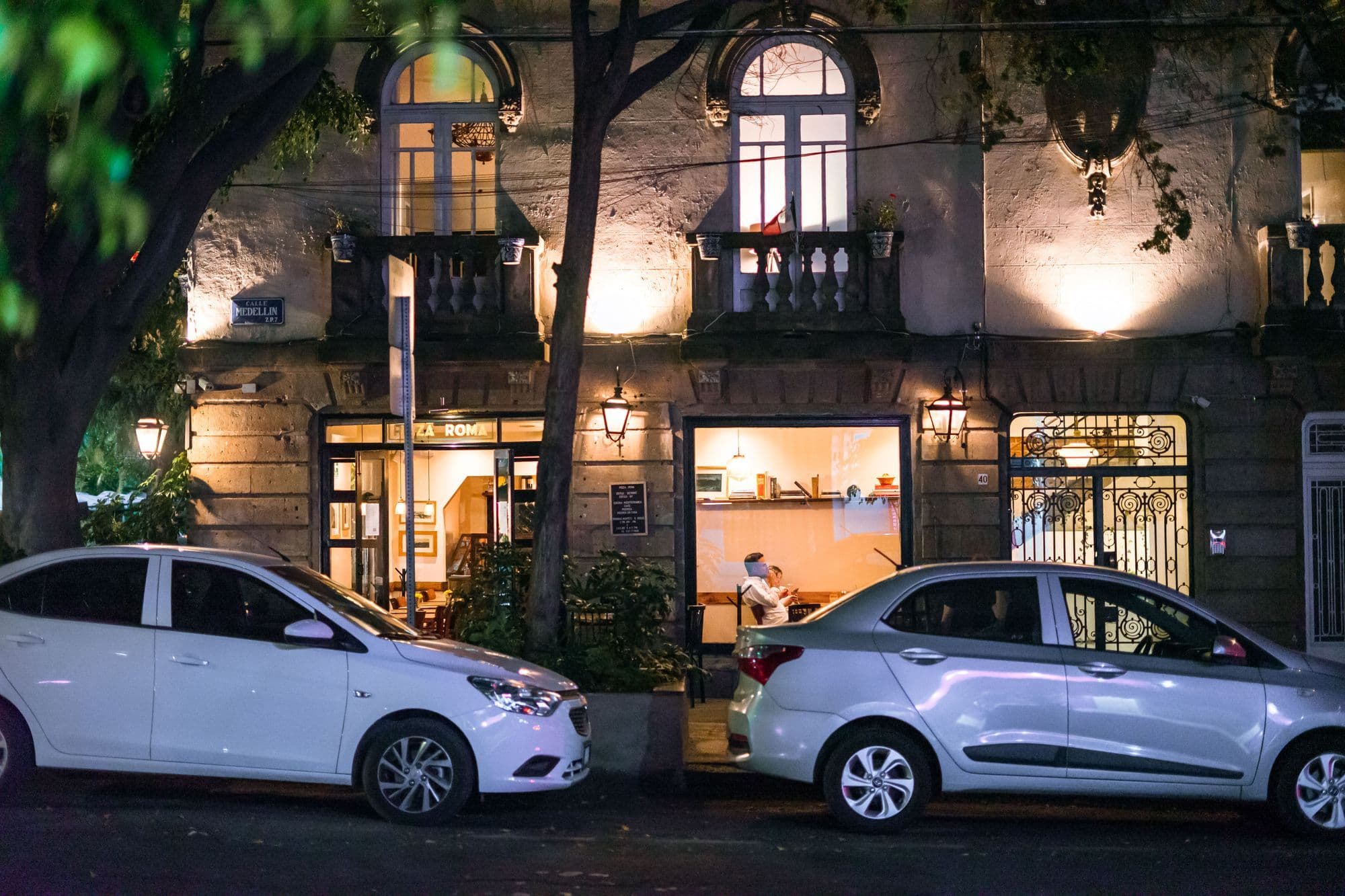 A lit café on the ground floor of an old stone building with balconies, two cars parked outside at night.