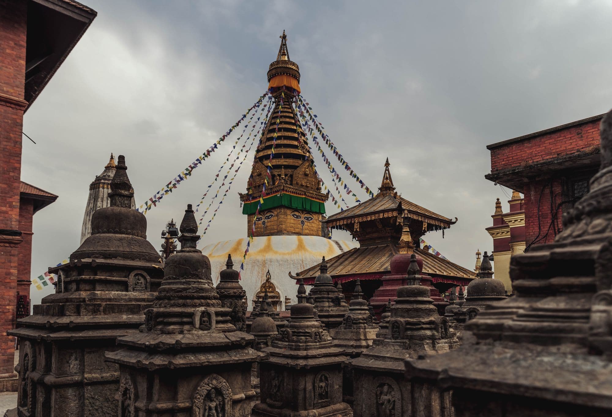 The white dome and painted eyes of Swayambhunath Stupa rising above smaller stone shrines on a cloudy day in Kathmandu.