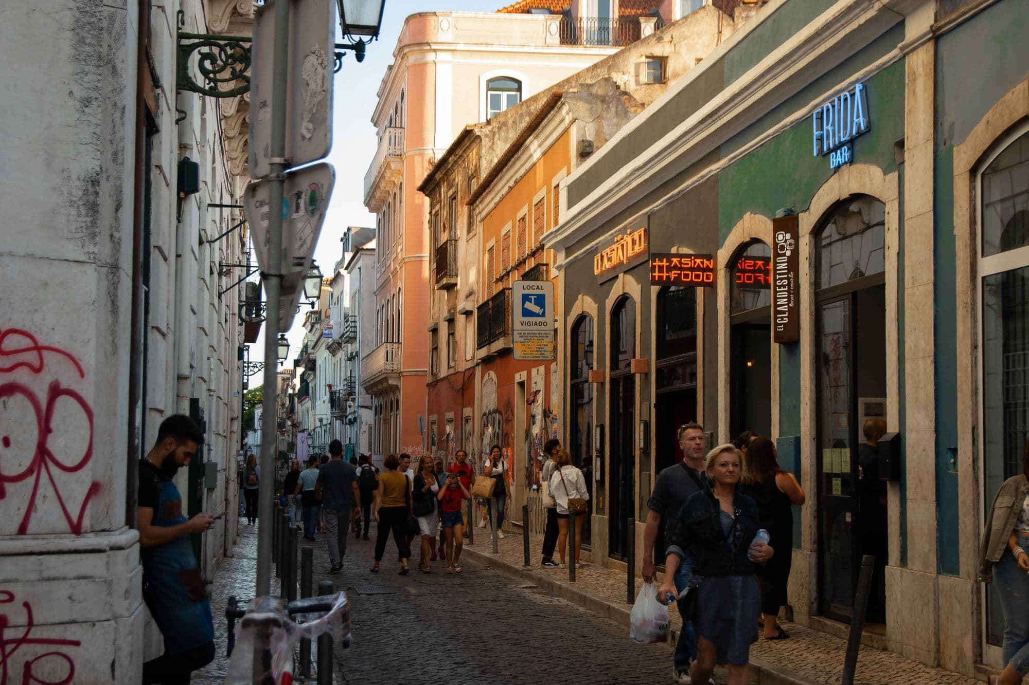 People walk down a narrow cobbled street lined with pastel facades and shopfronts in central Lisbon.