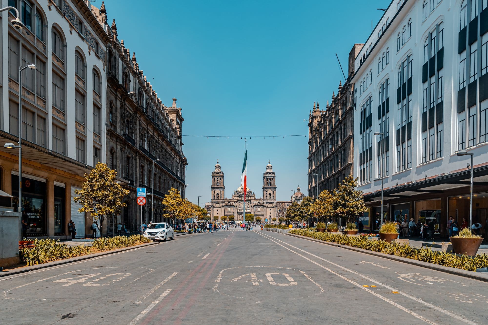 A wide, empty street in Mexico City's historic centre leads to the twin towers of the Metropolitan Cathedral, with a large Mexican flag in front.