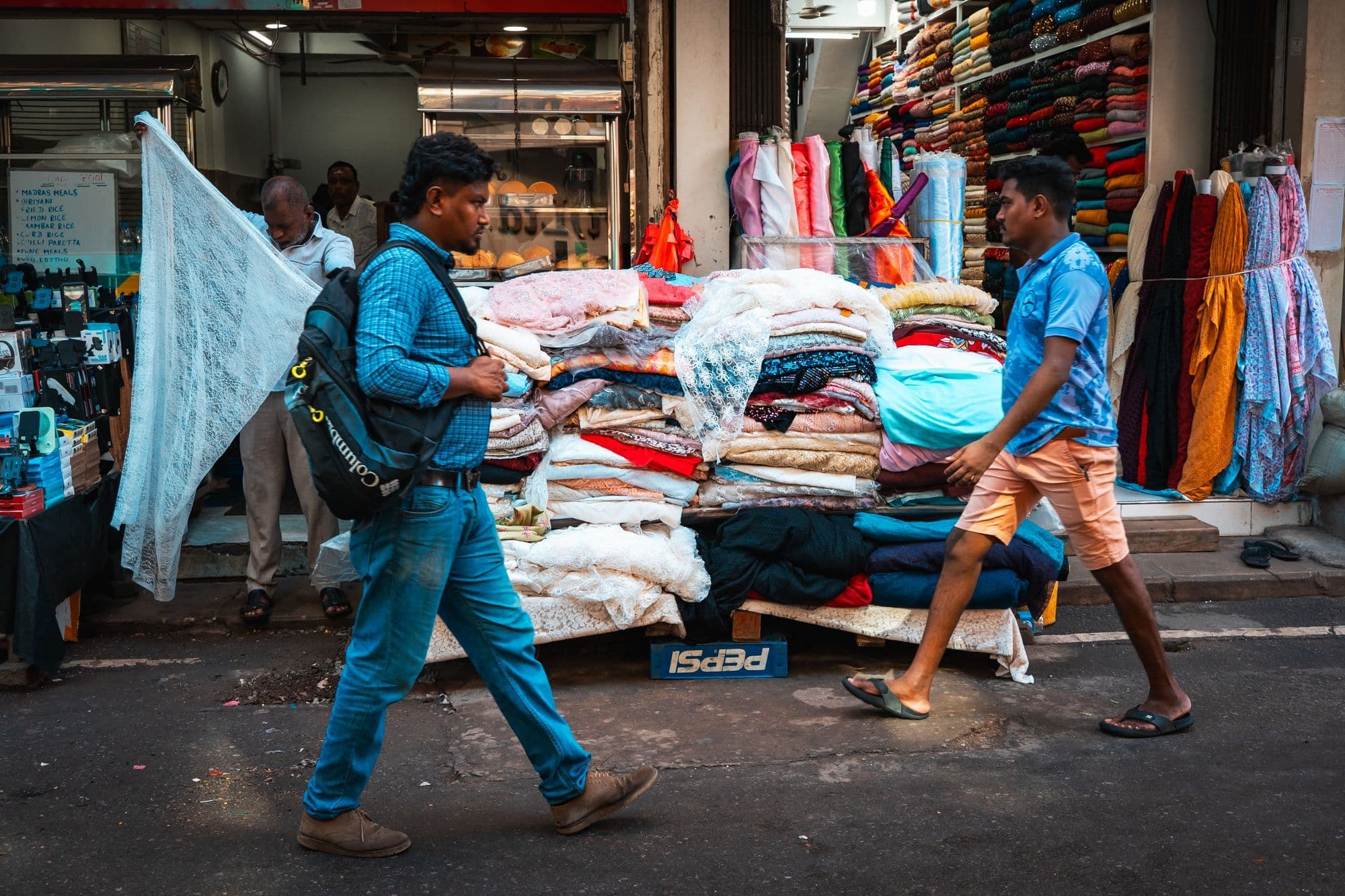 Two men walk past a street stall piled with folded textiles in Colombo's Pettah market.